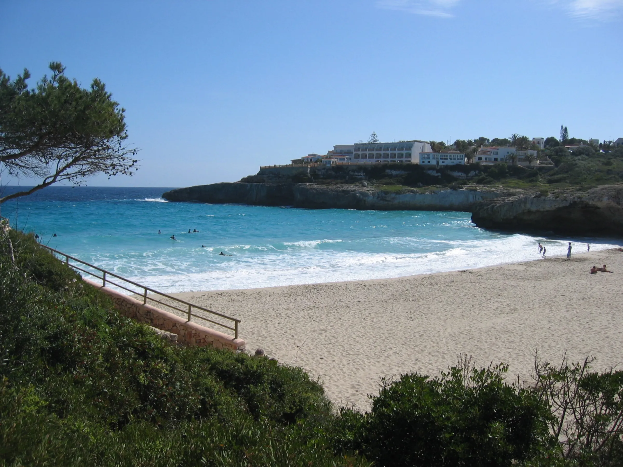 Cala Domingos desde lo alto, con la playa de arena blanca, el agua azul y la punta de roca con un hotel al fondo