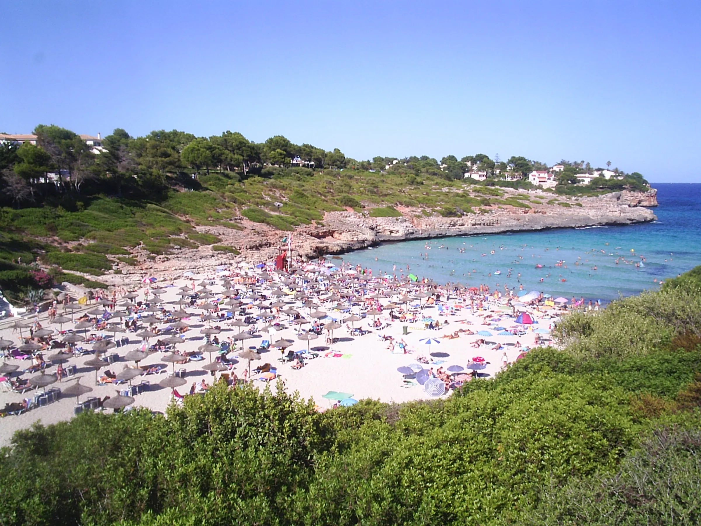 Cala Mendia desde lo alto en pleno verano, con la arena llena de sombrillas y el entrante turquesa entre laderas de matorral