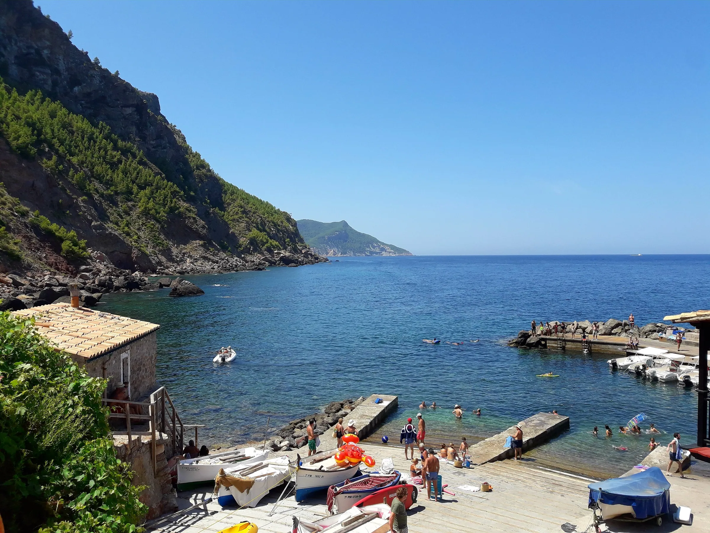 Bañistas en las plataformas y la rampa del Port de Valldemossa, con el agua azul abierta al mar y la costa de la Tramuntana al fondo