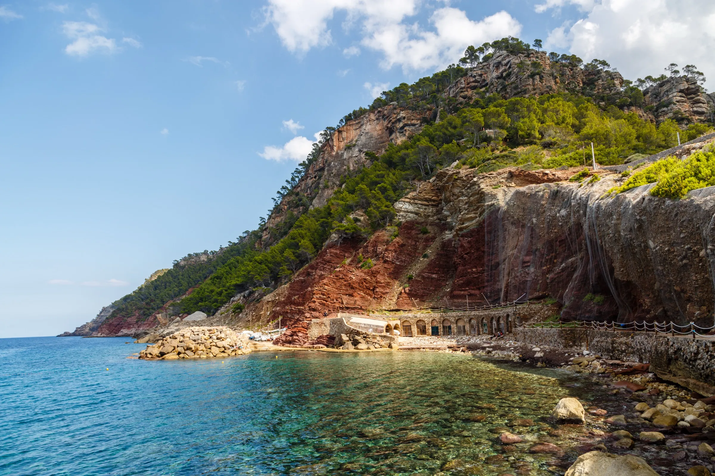 La orilla de cantos de Cala d'Estellencs con agua transparente, las casetas bajo el acantilado de tierra roja y la ladera de pinar