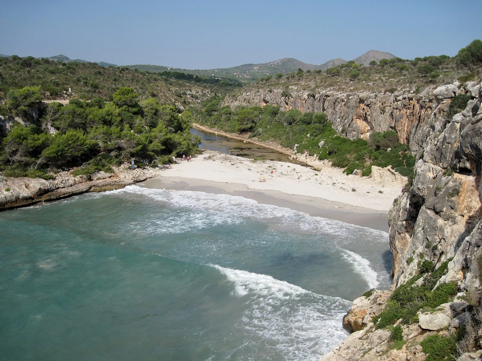 Cala Magraner vista desde el acantilado, con la arena blanca, el torrente al fondo y la pared de roca a la derecha