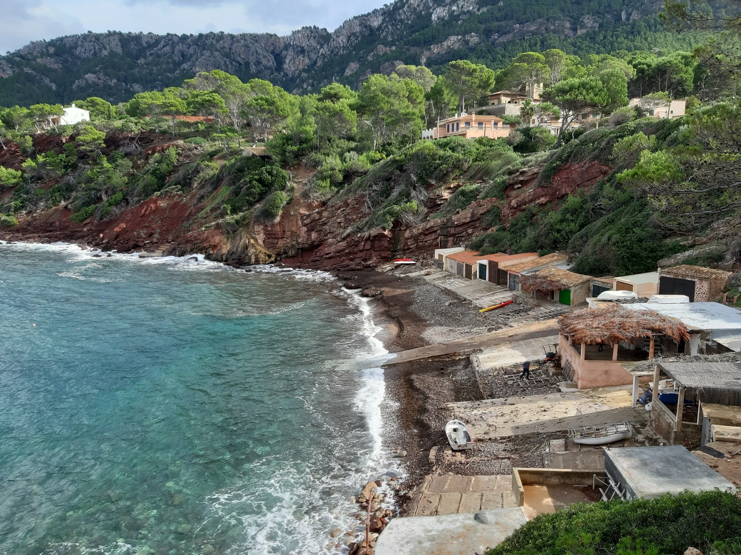 Vista desde lo alto del Port des Canonge, con la playa de cantos, el agua clara, la fila de casetas de pescadores y el acantilado rojo con pinar