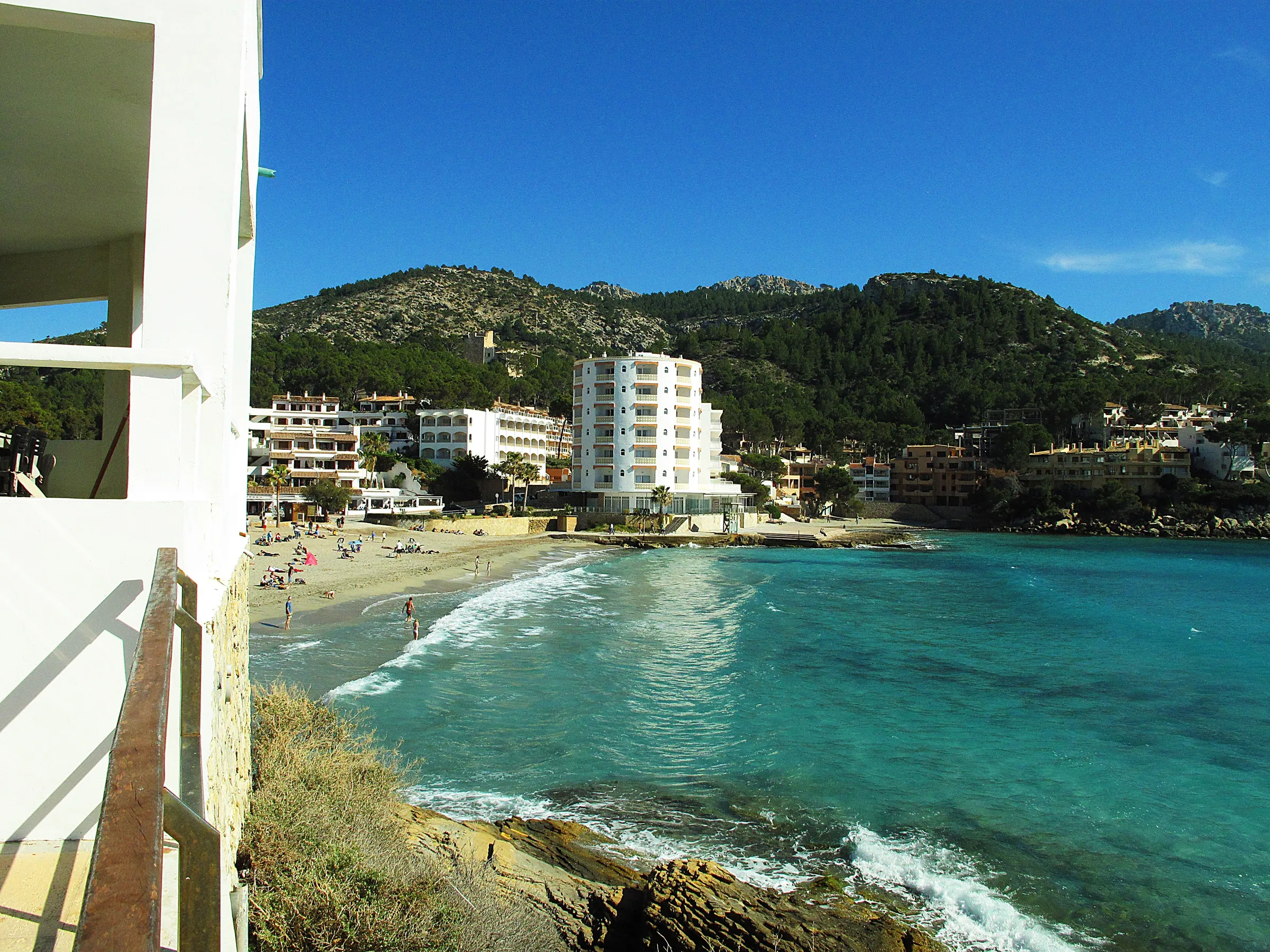 La playa de Sant Elm vista desde las rocas, con el agua turquesa, la arena y el pueblo al pie de la montaña