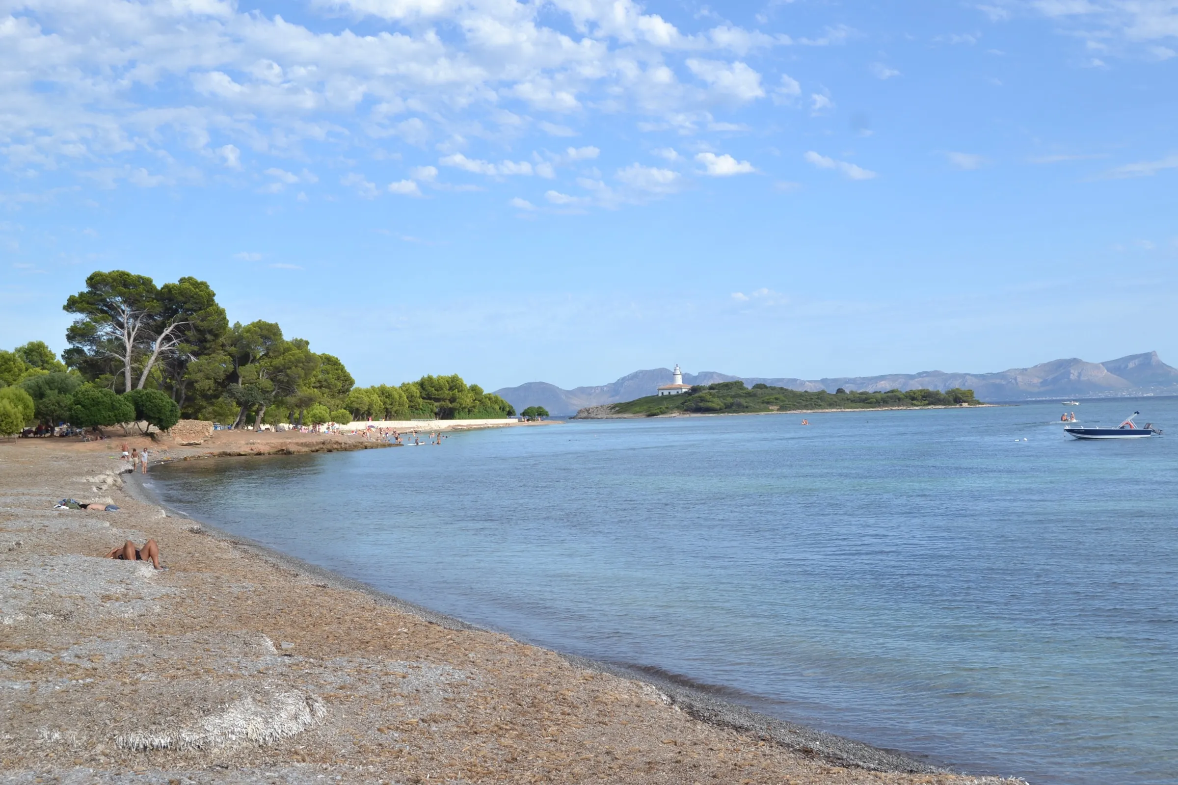 Alcanada beach, with pebbles at the water's edge, the pine wood and the islet with its lighthouse behind