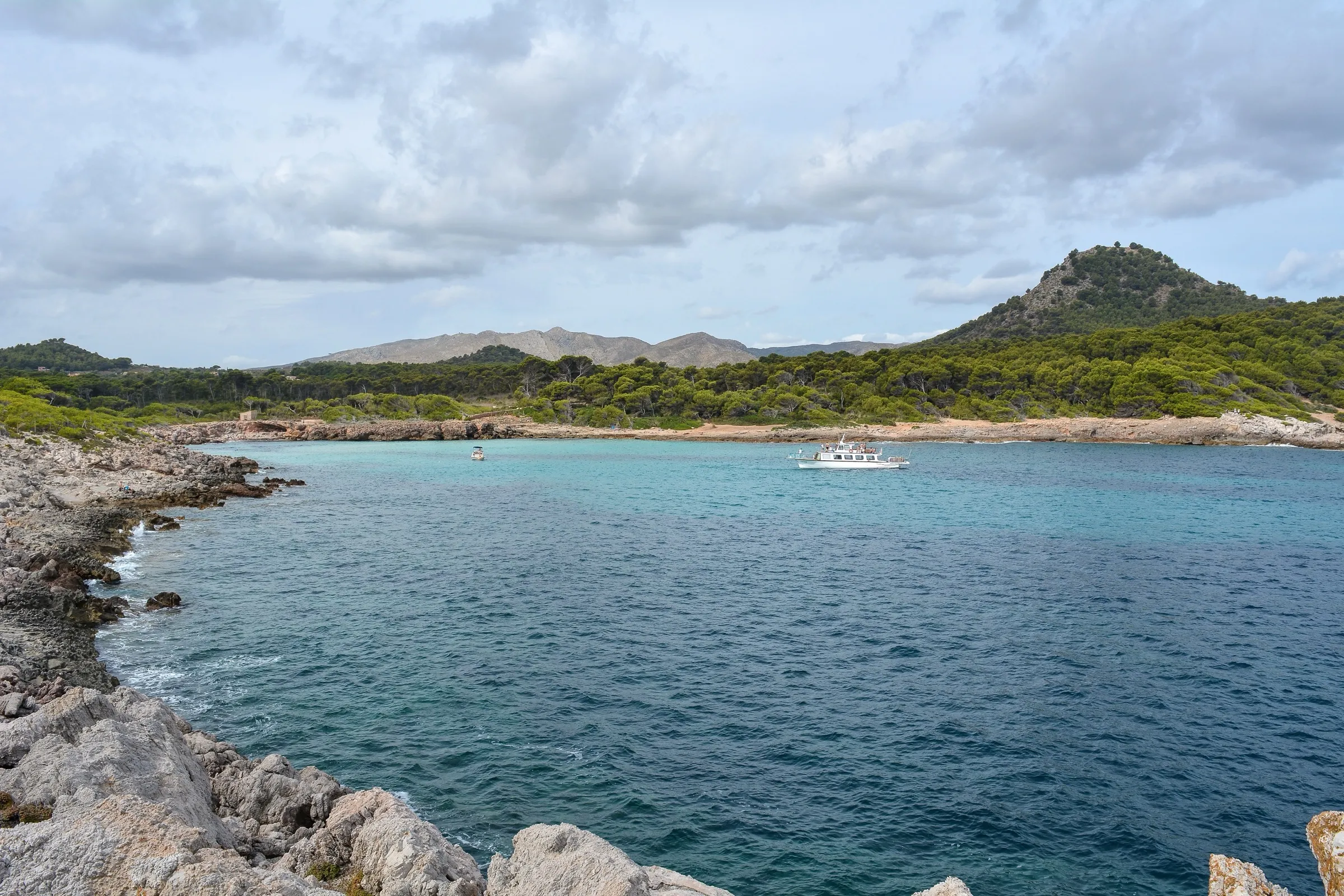 Cala Moltó desde las rocas de la punta, con el agua azul, una barca fondeada y el pinar y la montaña al fondo