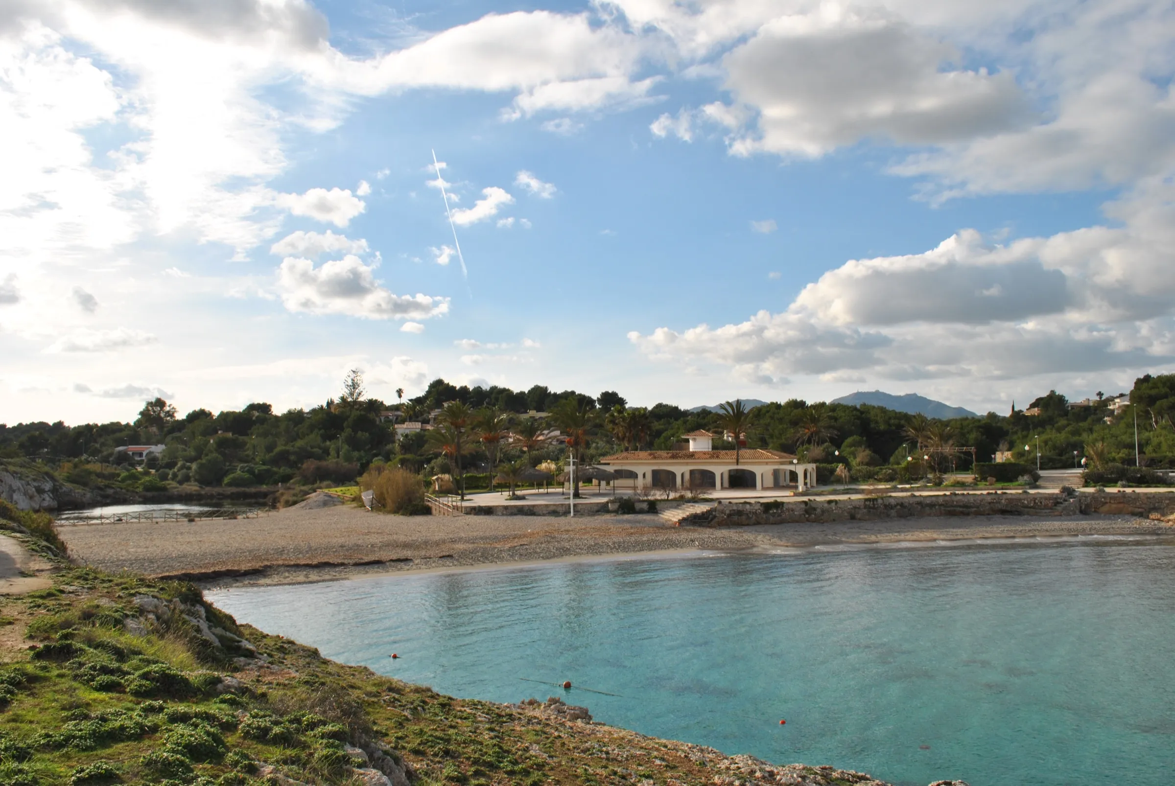 El agua turquesa de Cala Murada desde el lateral, con la playa de arena y un edificio de arcos al fondo
