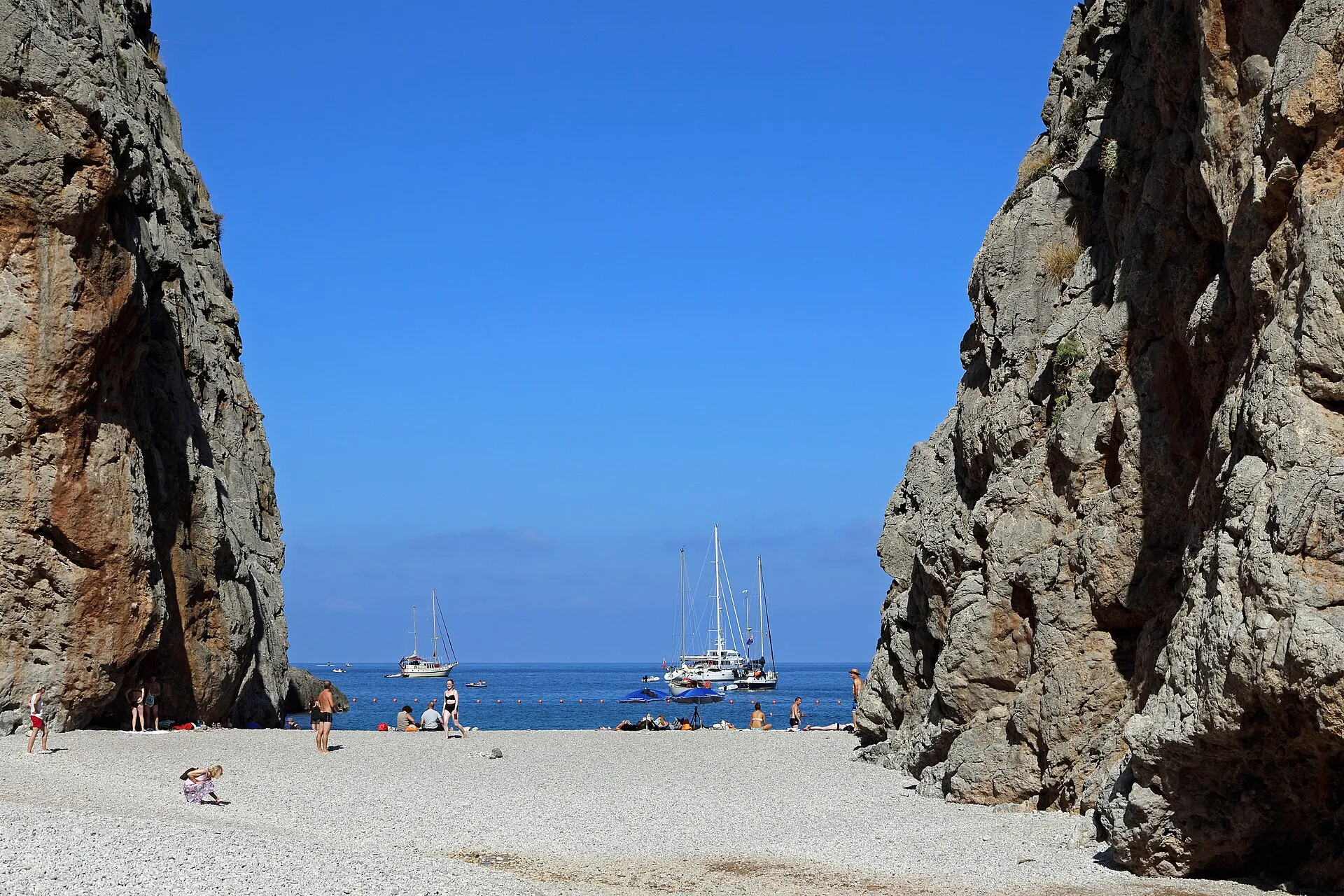 La playa de cantos del Torrent de Pareis entre dos paredes de roca, con el mar azul y veleros fondeados al fondo