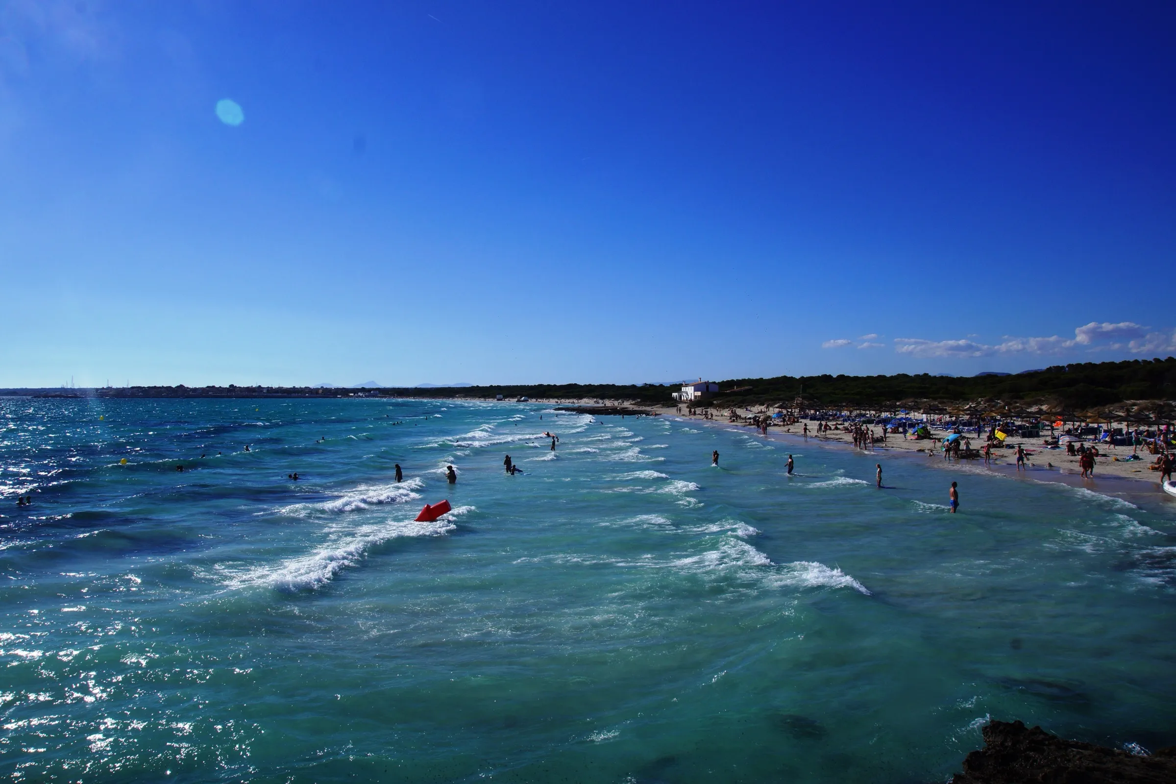 Arenal de sa Ràpita en verano, con olas suaves, agua turquesa y bañistas a lo largo de la orilla
