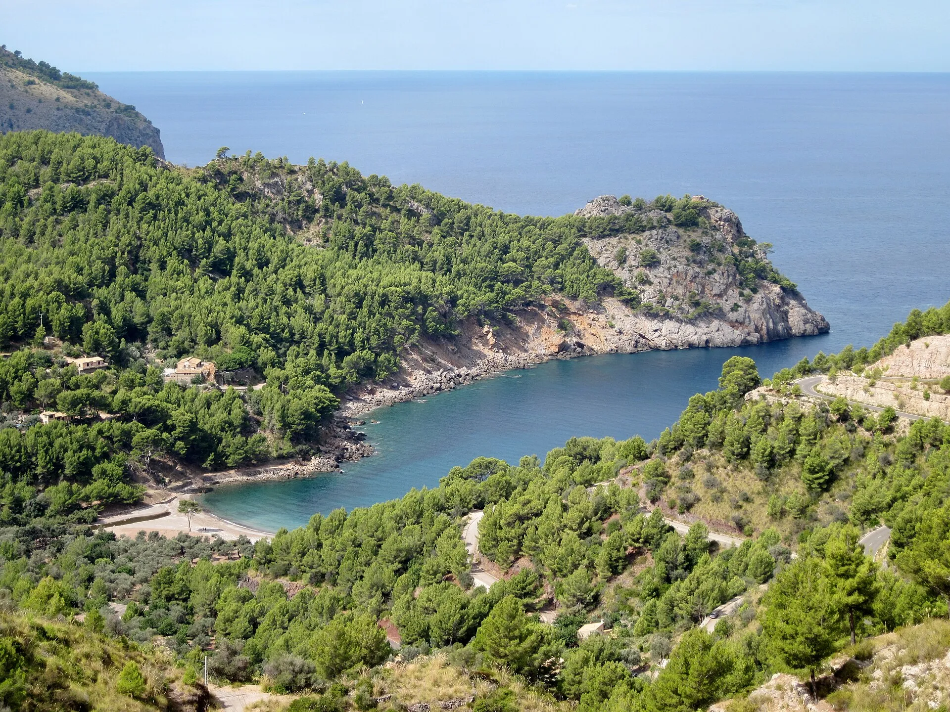 Vista desde lo alto de Cala Tuent, con el agua azul encajada entre laderas de pinar y un cabo de roca que la cierra hacia el mar