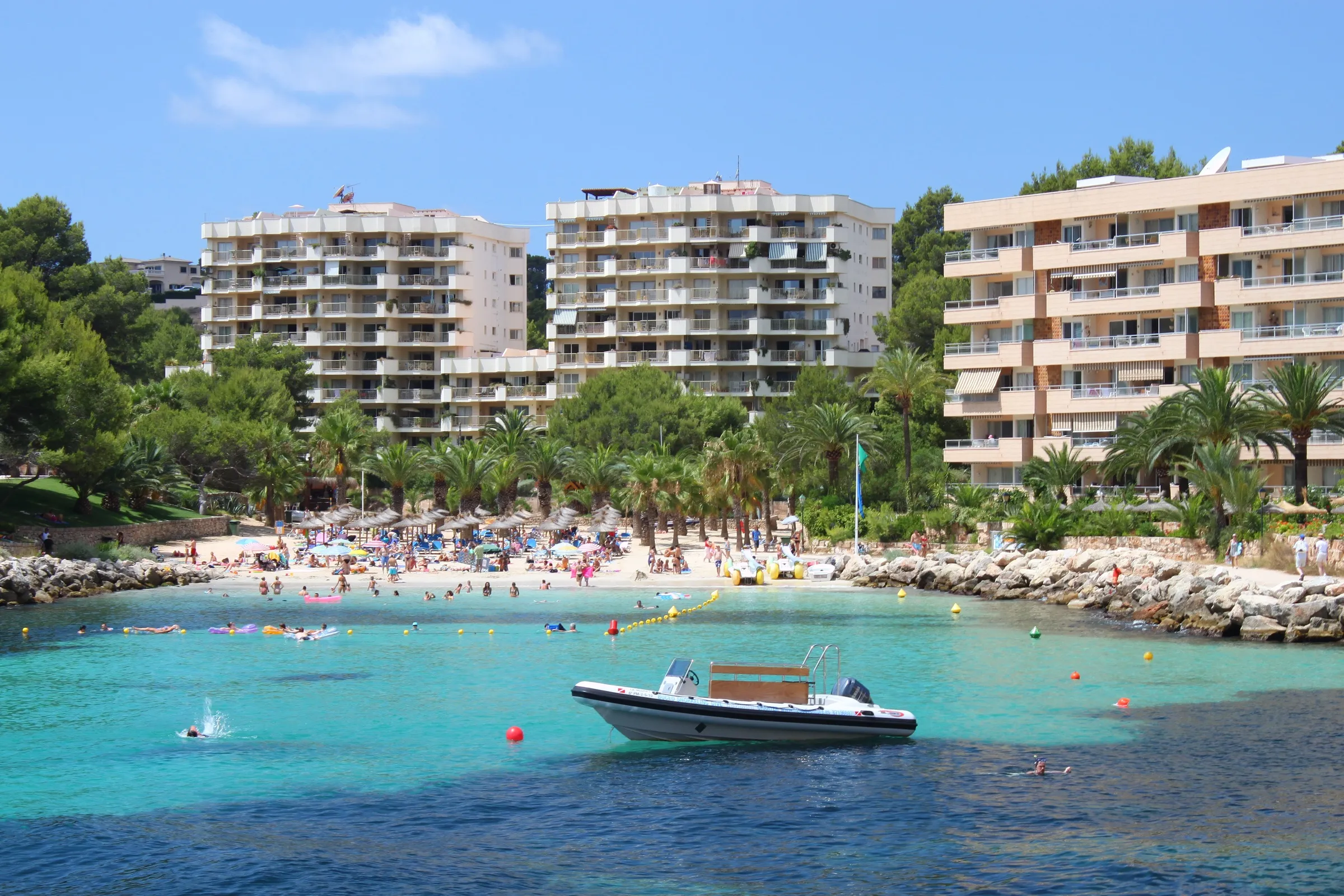 Cala Vinyes en verano, con el agua turquesa, la arena llena de bañistas, palmeras y edificios de apartamentos detrás