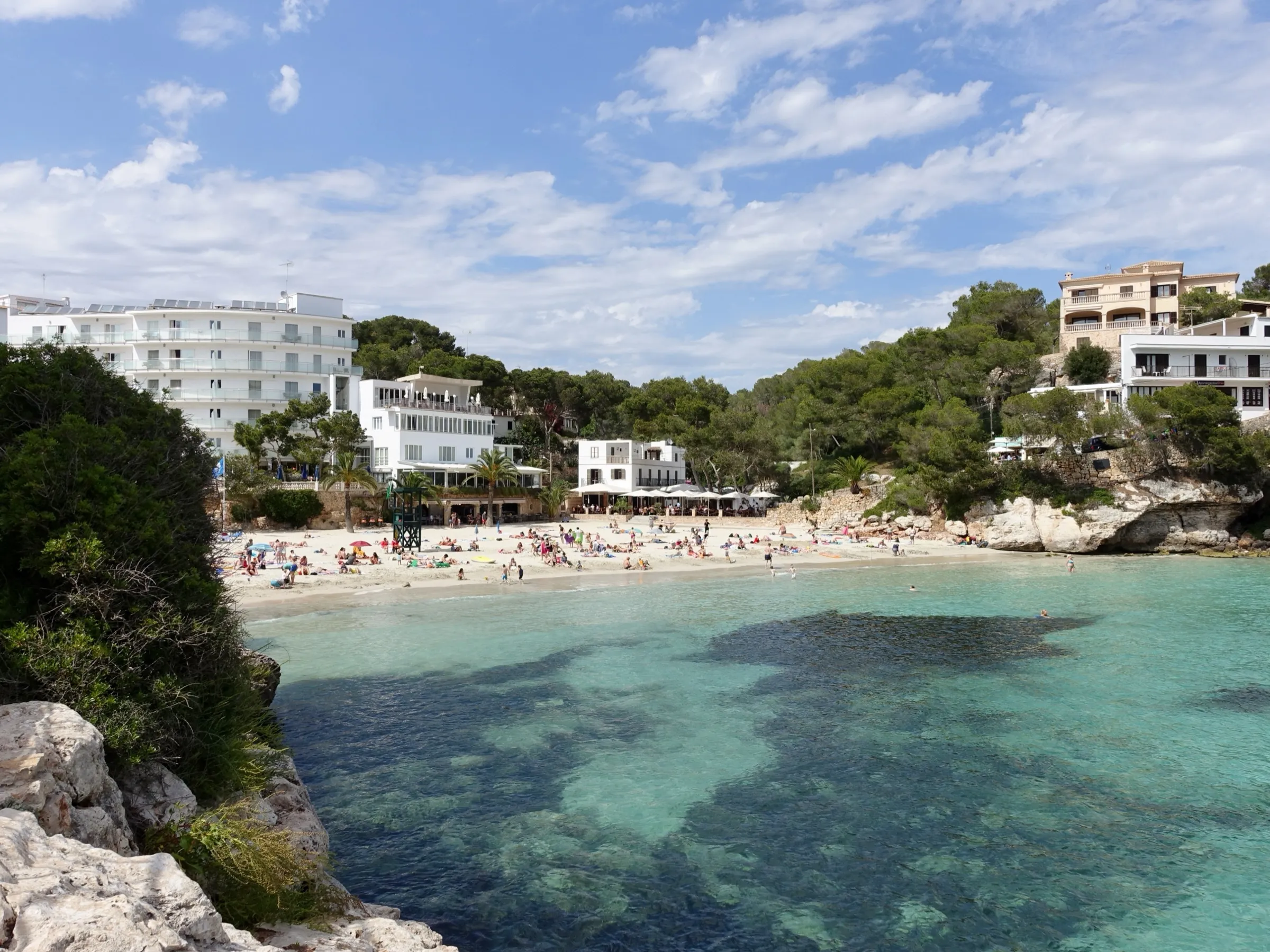 Cala Santanyí vista desde las rocas, con el agua turquesa, la playa de arena blanca y los hoteles entre pinos al fondo