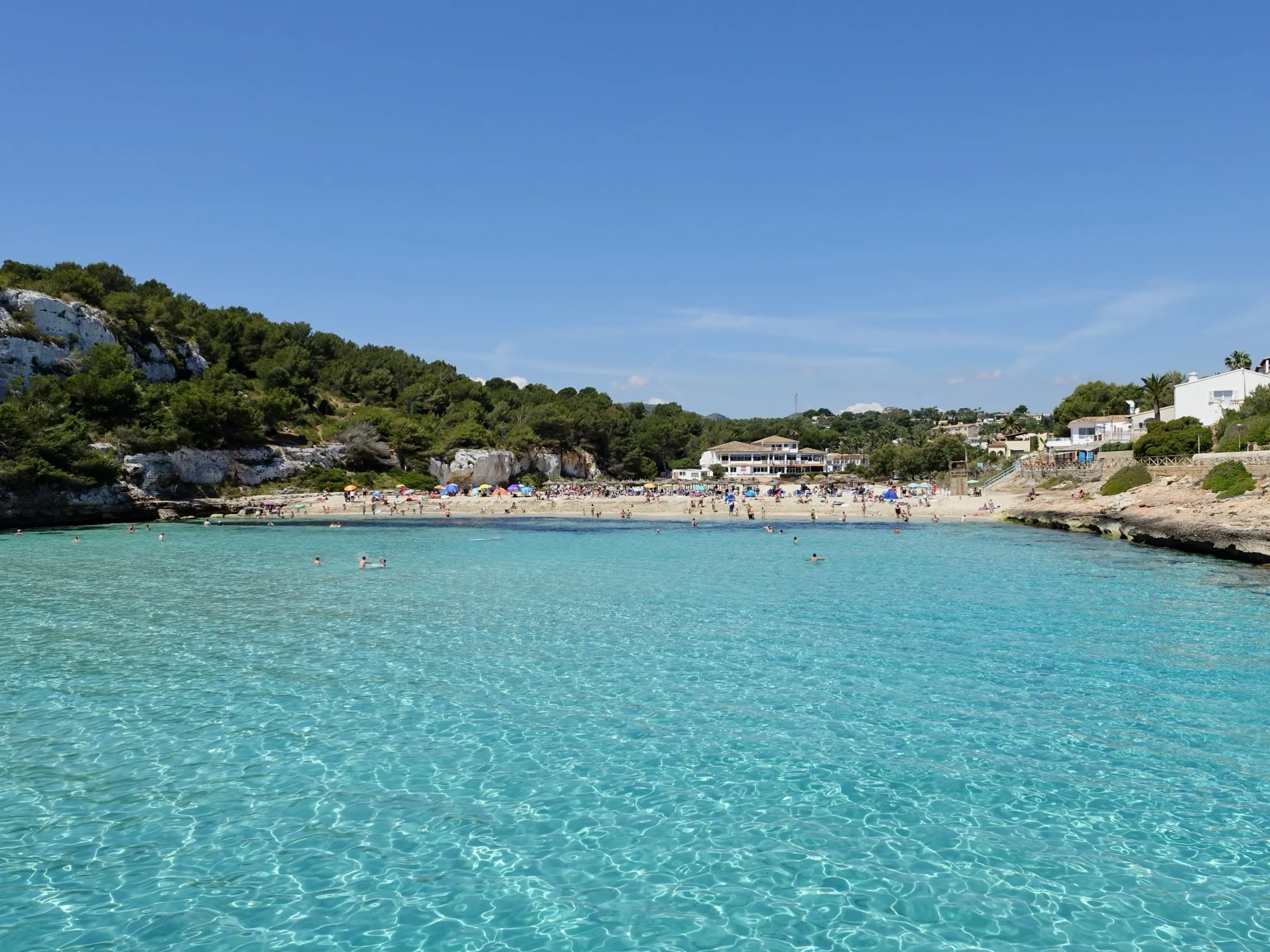 La playa de s'Estany d'en Mas al fondo de la cala, con el agua turquesa y transparente en primer plano y pinar a los lados