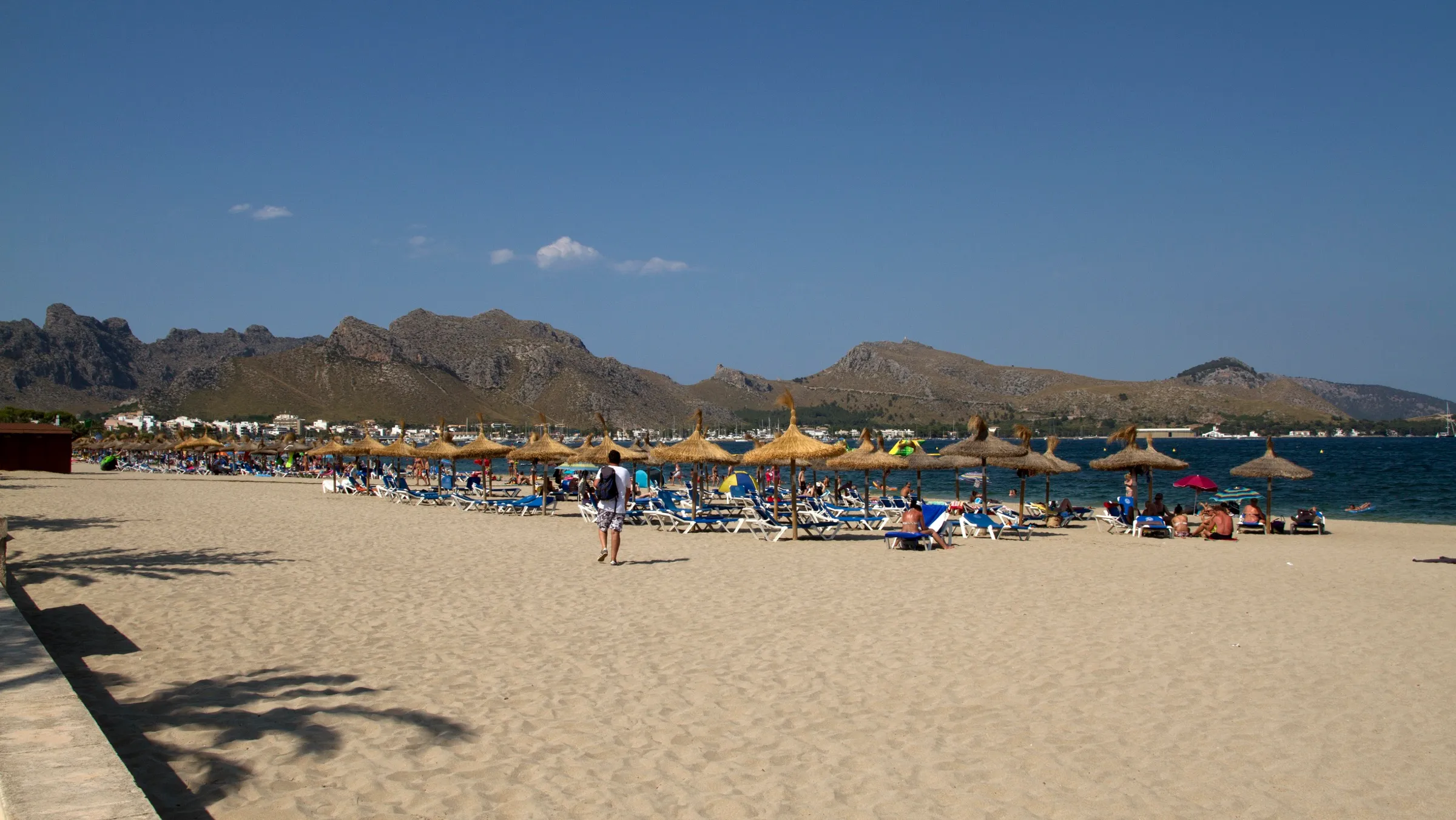 The sand at Llenaire with straw parasols and sunbeds, and the Serra de Tramuntana mountains in the distance