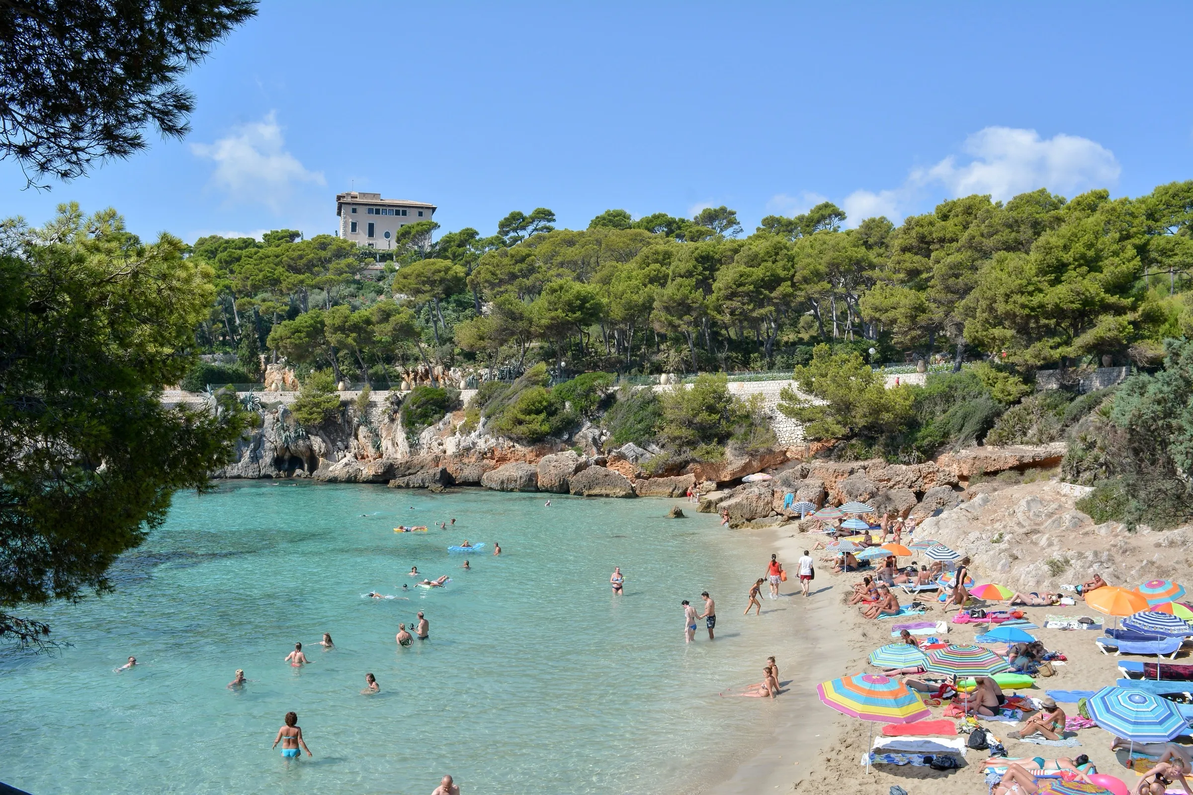 Cala Gat con bañistas en el agua turquesa y la arena, rodeada de pinar y con la casa de Juan March en lo alto