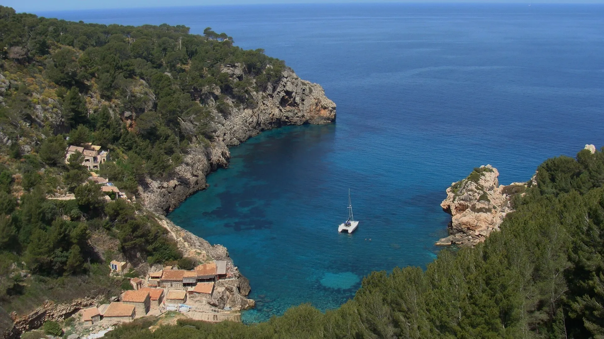Vista desde lo alto de Cala Deià, con el agua azul turquesa entre acantilados con pinar y las casetas de pescadores abajo