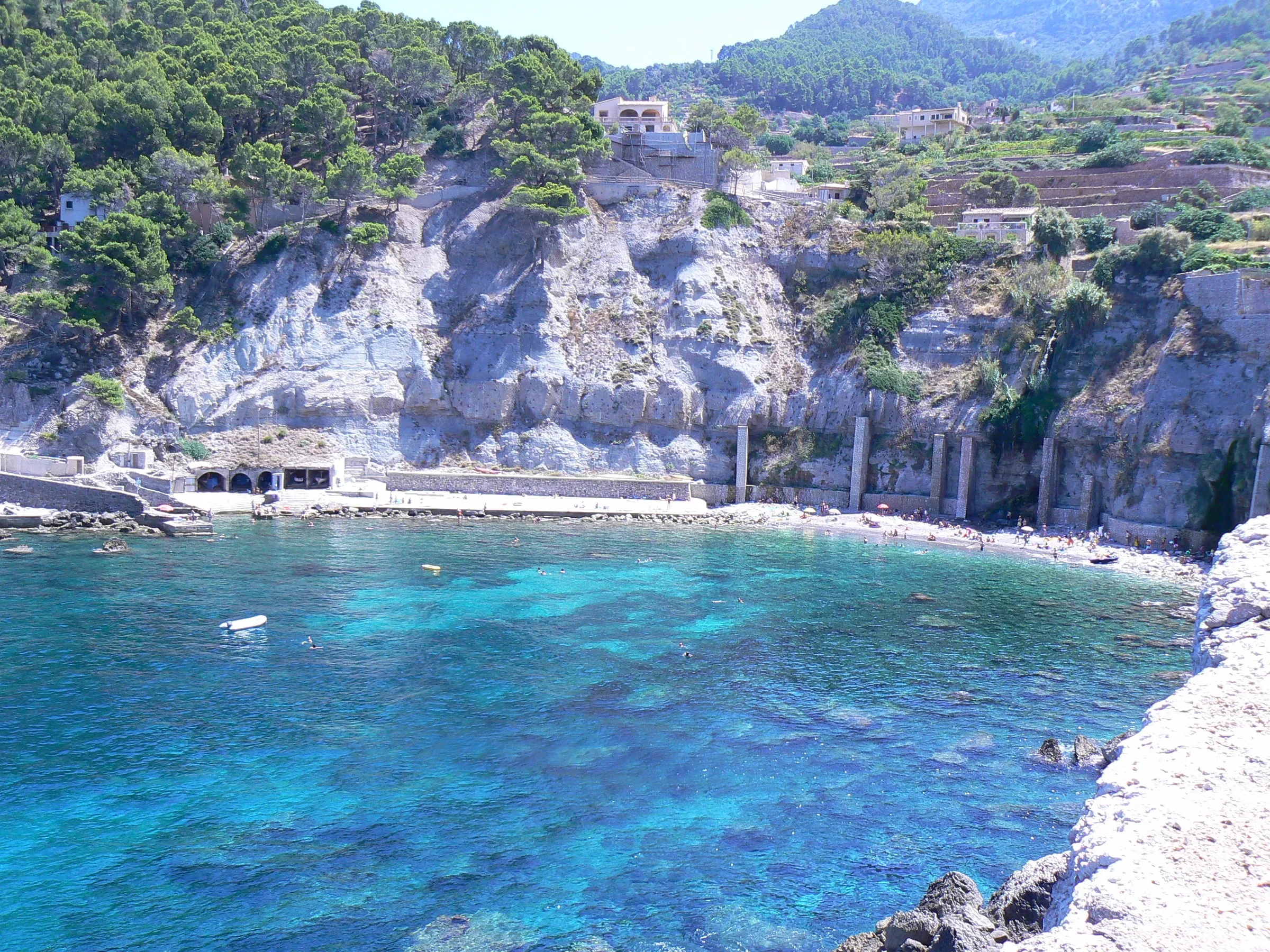 View from above the Port de Banyalbufar, with the turquoise water, the huts and the walkway at the foot of the white cliff