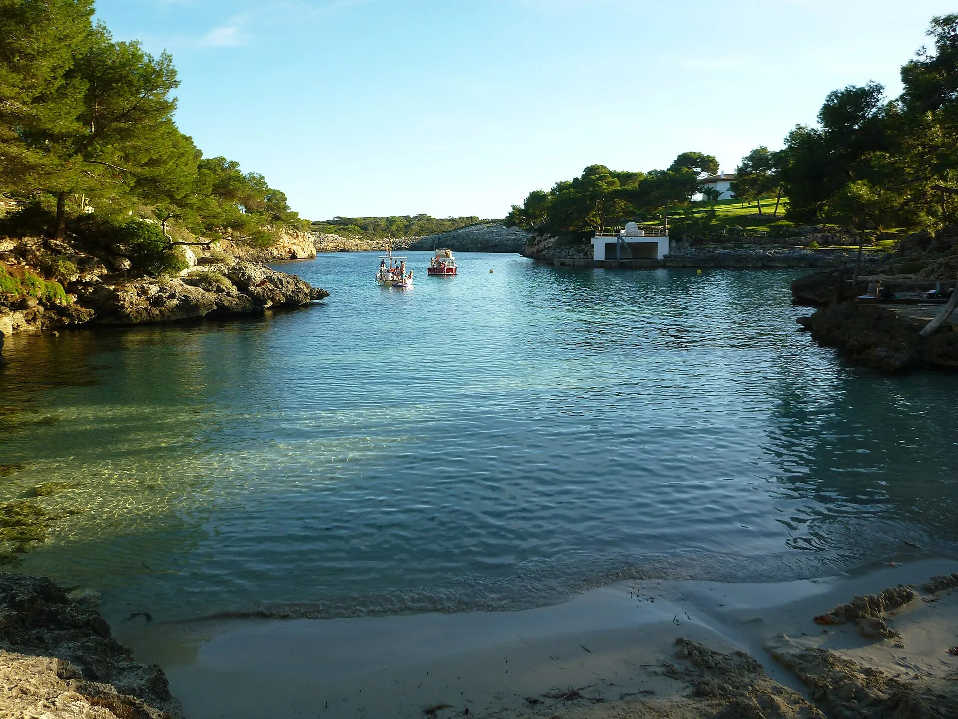 Cala Mitjana vista desde la arena, con el brazo de mar entre rocas y pinos y dos barcas fondeadas al fondo