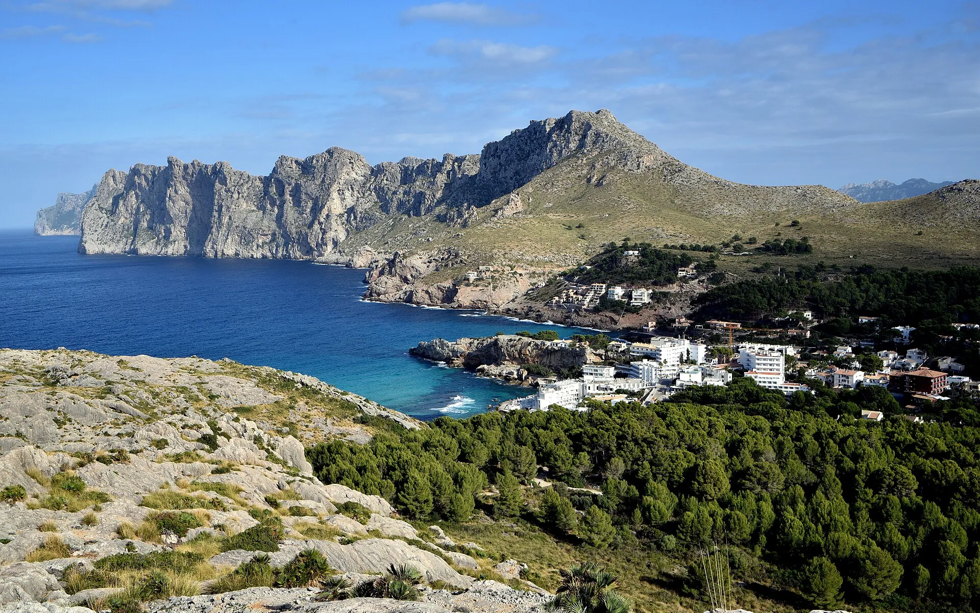 Vista desde lo alto de la bahía de Cala Sant Vicenç, con el pueblo blanco junto al mar, el pinar y la cresta del Cavall Bernat al fondo