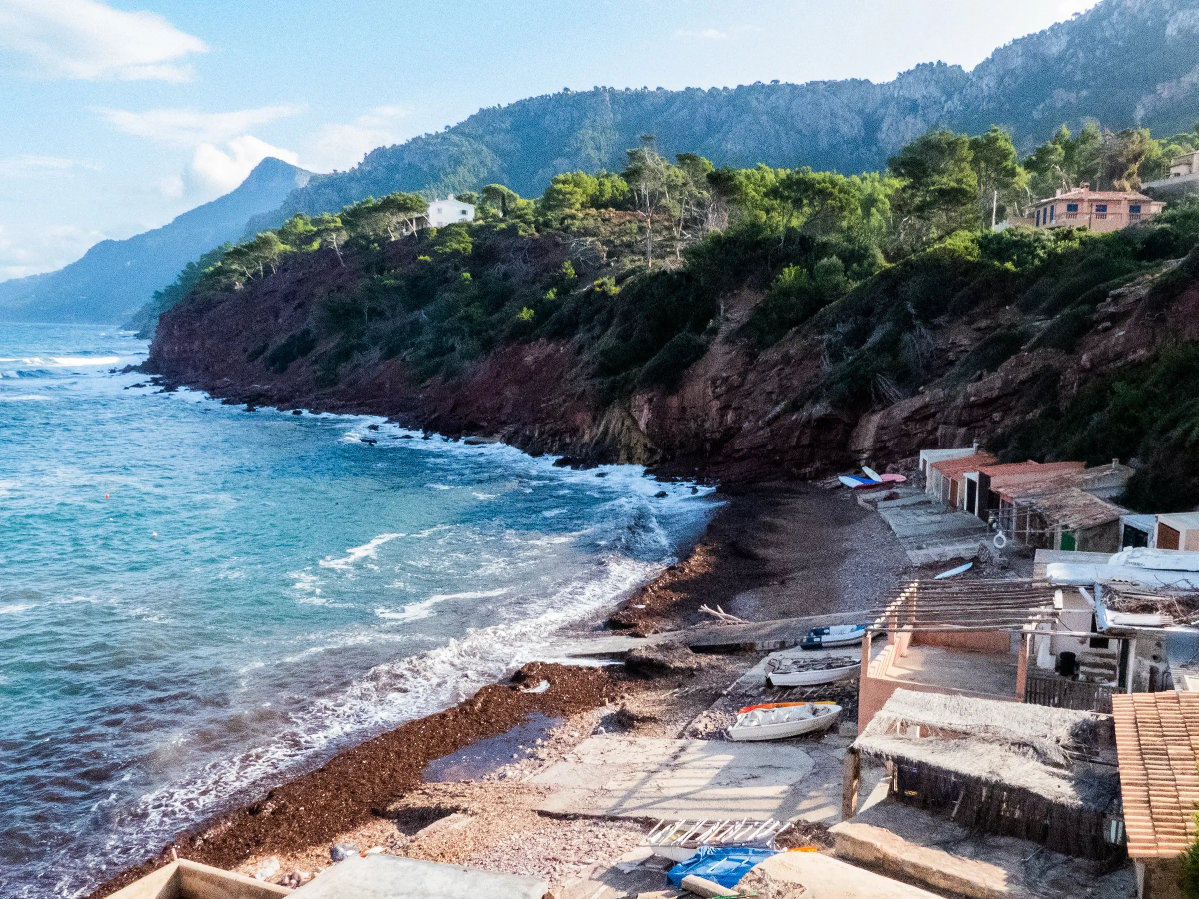 La playa del Port des Canonge con barcas varadas junto a las casetas y la costa de la Serra de Tramuntana al fondo