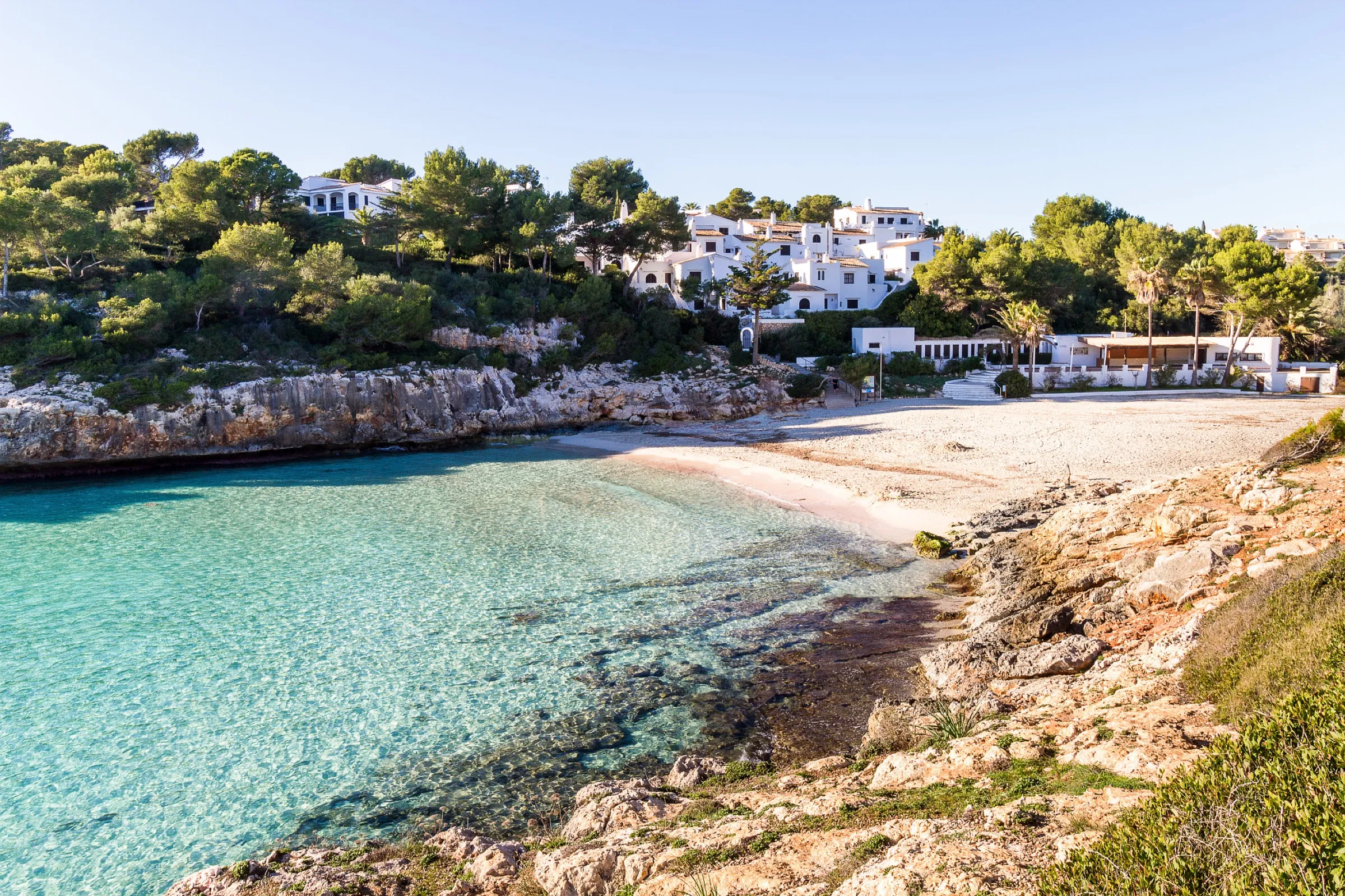 La playa de arena de Cala Anguila y el agua transparente, con las casas blancas de la urbanización entre pinos