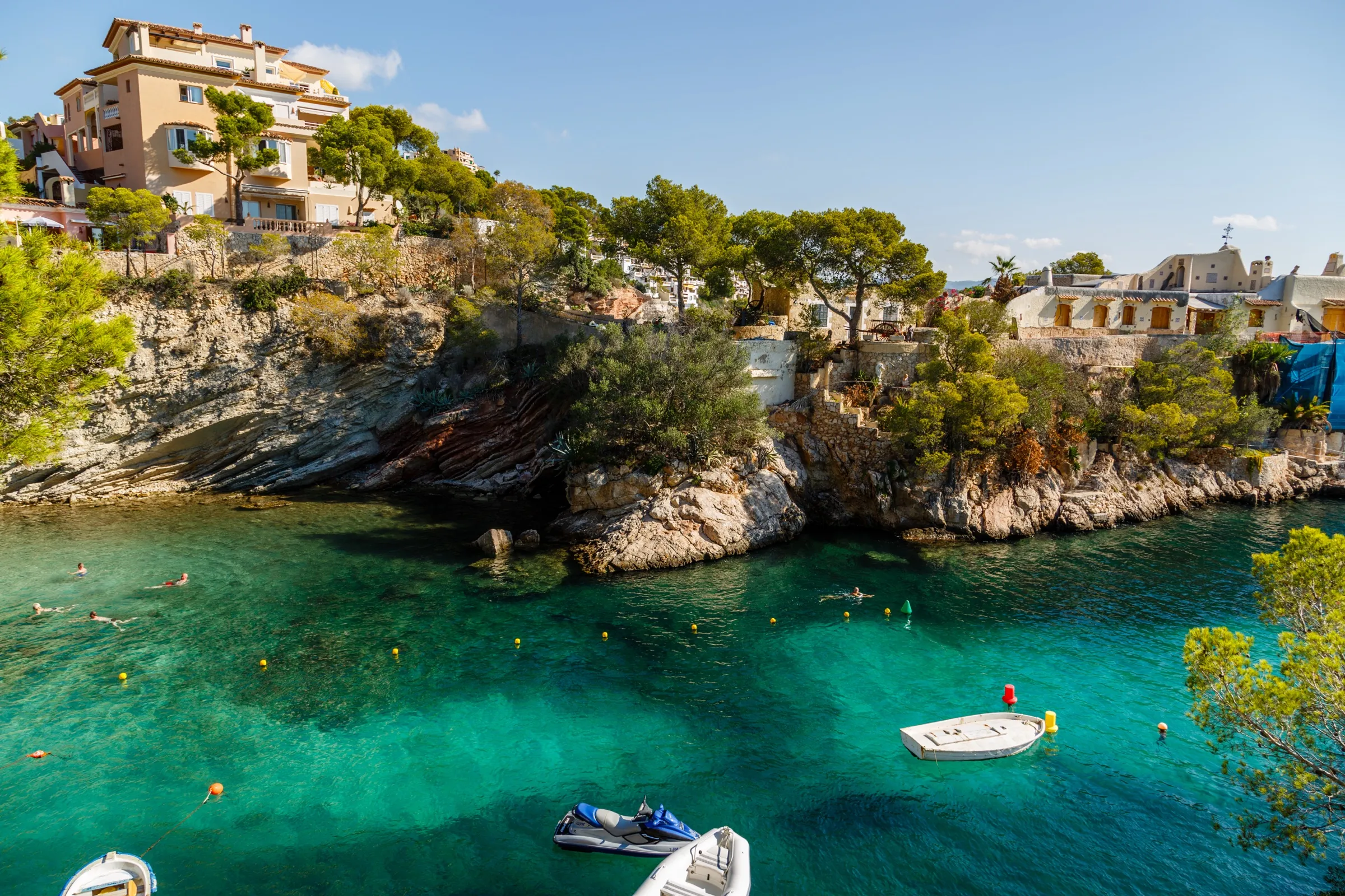 Agua turquesa junto a las rocas y el pinar de Cala Fornells, con barcas pequeñas y bañistas