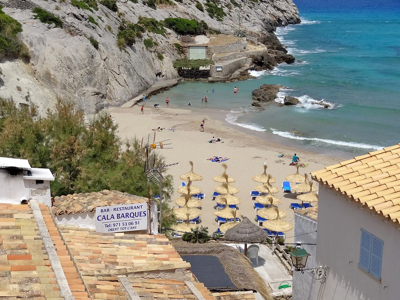 Cala Barques desde el pueblo, con sombrillas de paja en la arena, los antiguos varaderos de barcas y la pared de roca