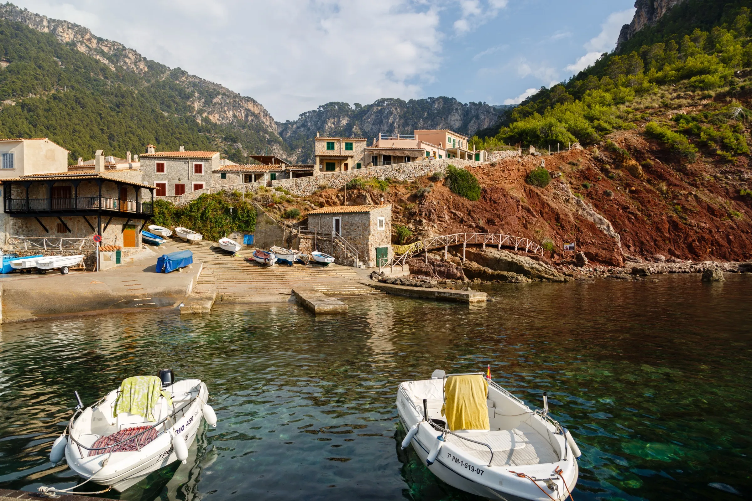 Barcas amarradas en el agua transparente del pequeño puerto de Valldemossa, con las casas y la ladera de tierra roja detrás
