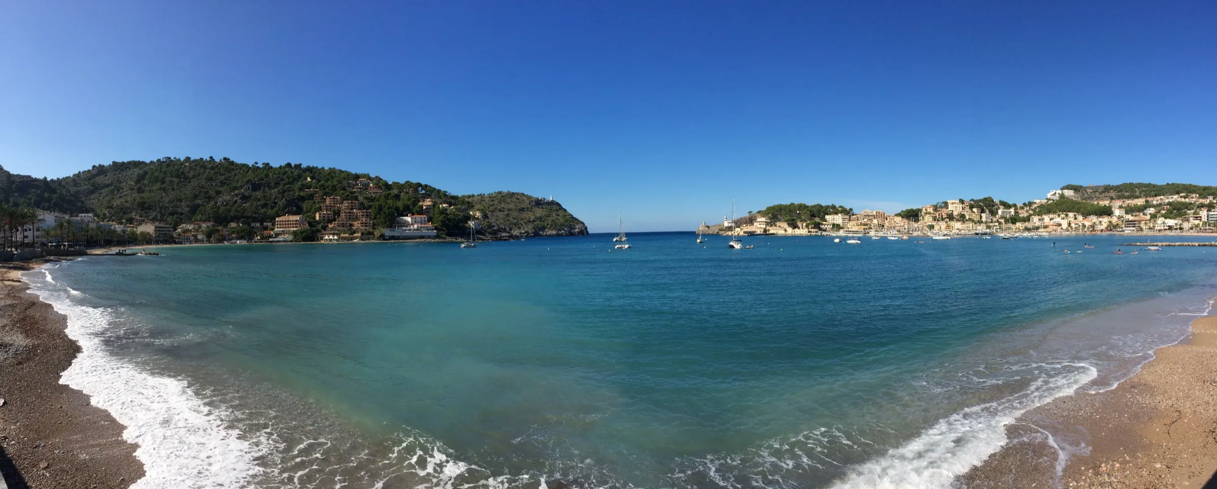 Panorámica de la bahía del Port de Sóller desde la playa, con el agua turquesa, olas suaves en la orilla y el pueblo a la derecha