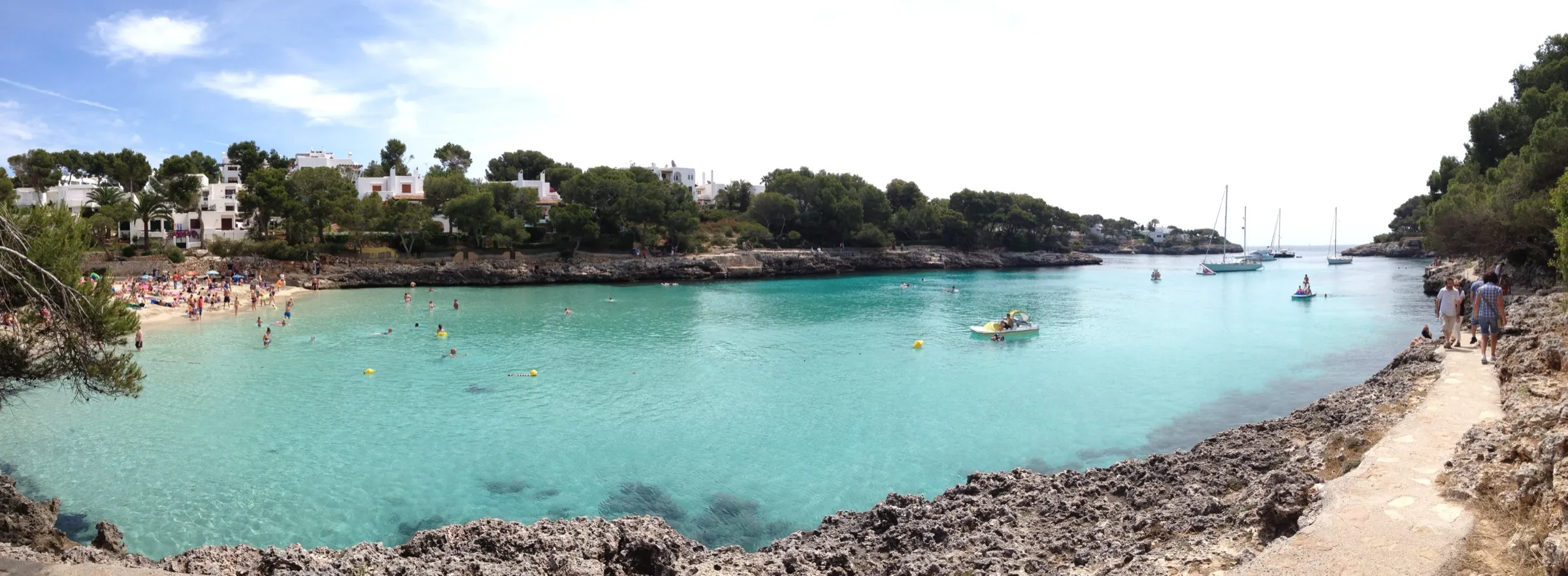 Panorama der Cala Gran mit türkisem Wasser, dem Strand links und dem felsigen Ufer im Vordergrund