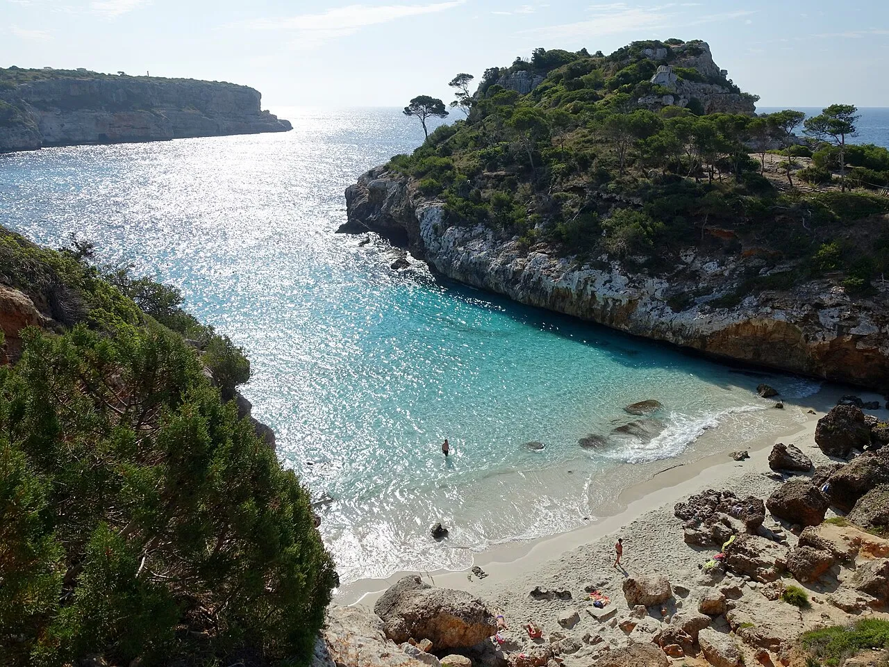 La playa del Caló des Moro con rocas en la orilla y el promontorio con pinos que cierra la cala
