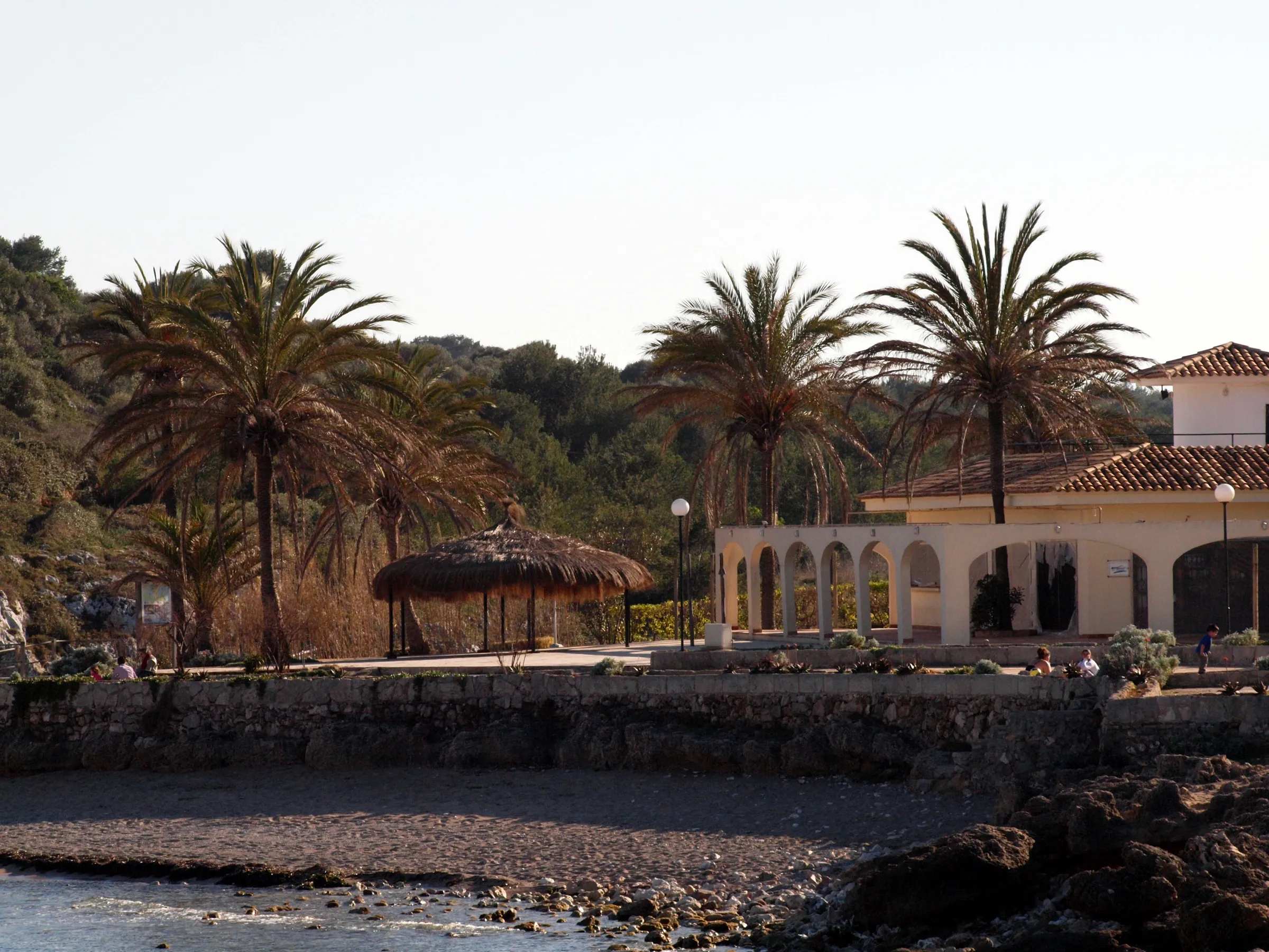 El paseo de Cala Murada con palmeras, una sombrilla de paja y el edificio de arcos sobre la orilla