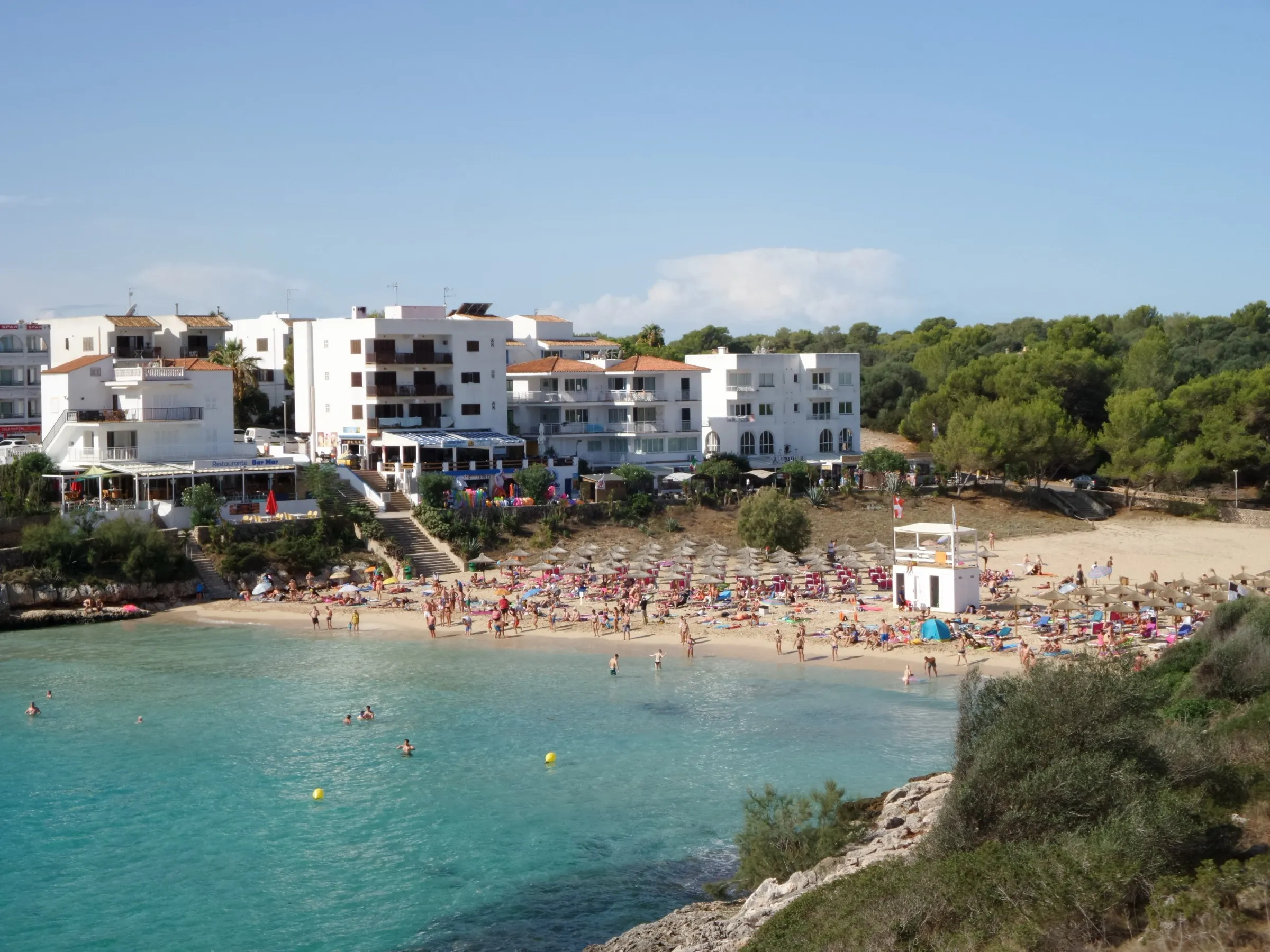 La playa de Cala Marçal llena de bañistas y sombrillas, con la torre del socorrista y los apartamentos blancos detrás