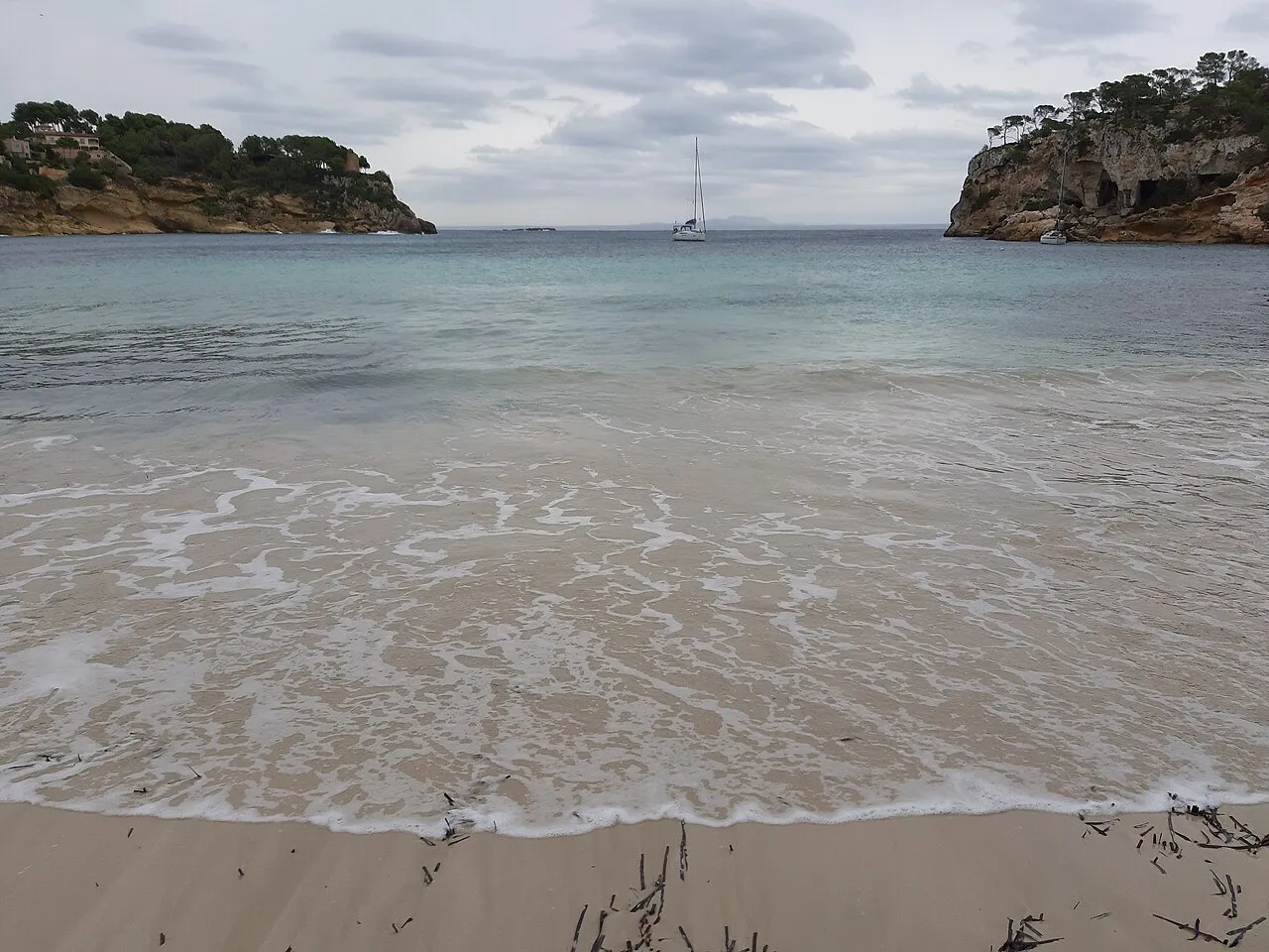 La orilla de Cala Portals Vells mirando hacia la boca de la ensenada, con un velero fondeado