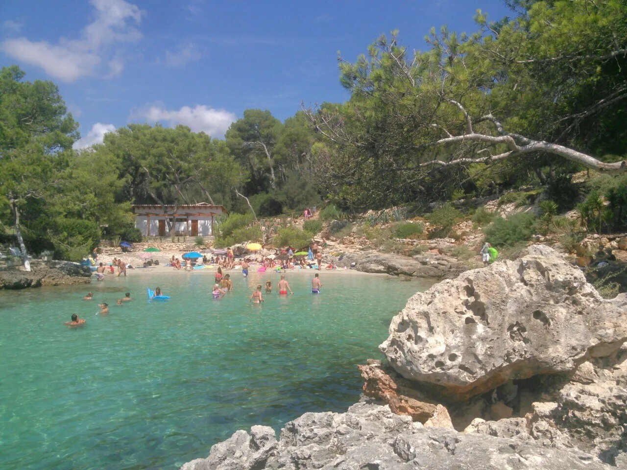 Bañistas en la pequeña playa de Cala Mitjana en verano, con agua turquesa, pinos y una caseta blanca detrás