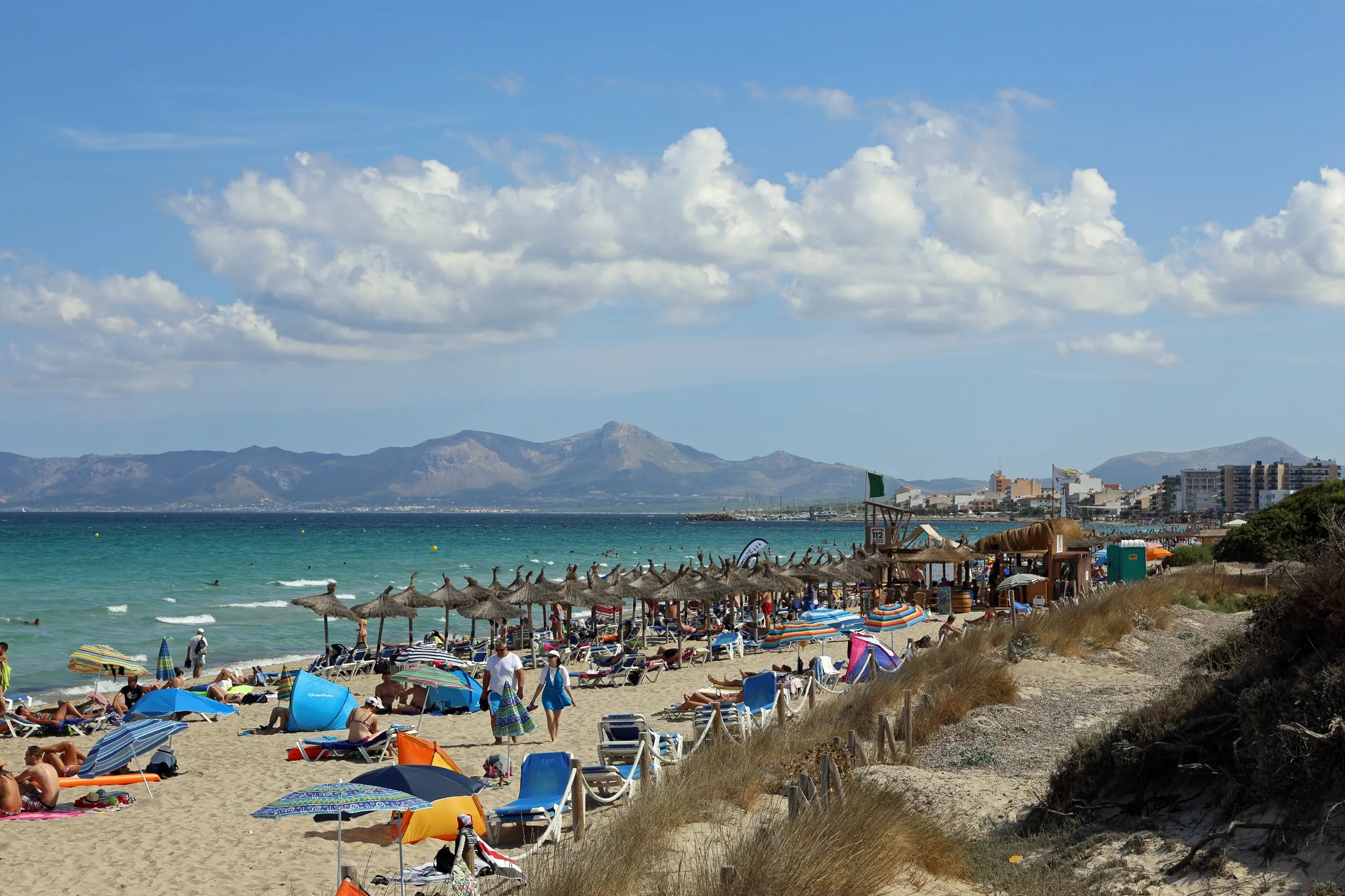 Strohschirme und Badegäste im Nordwestabschnitt des Strandes, rechts die Dünen, im Hintergrund die Berge von Alcúdia