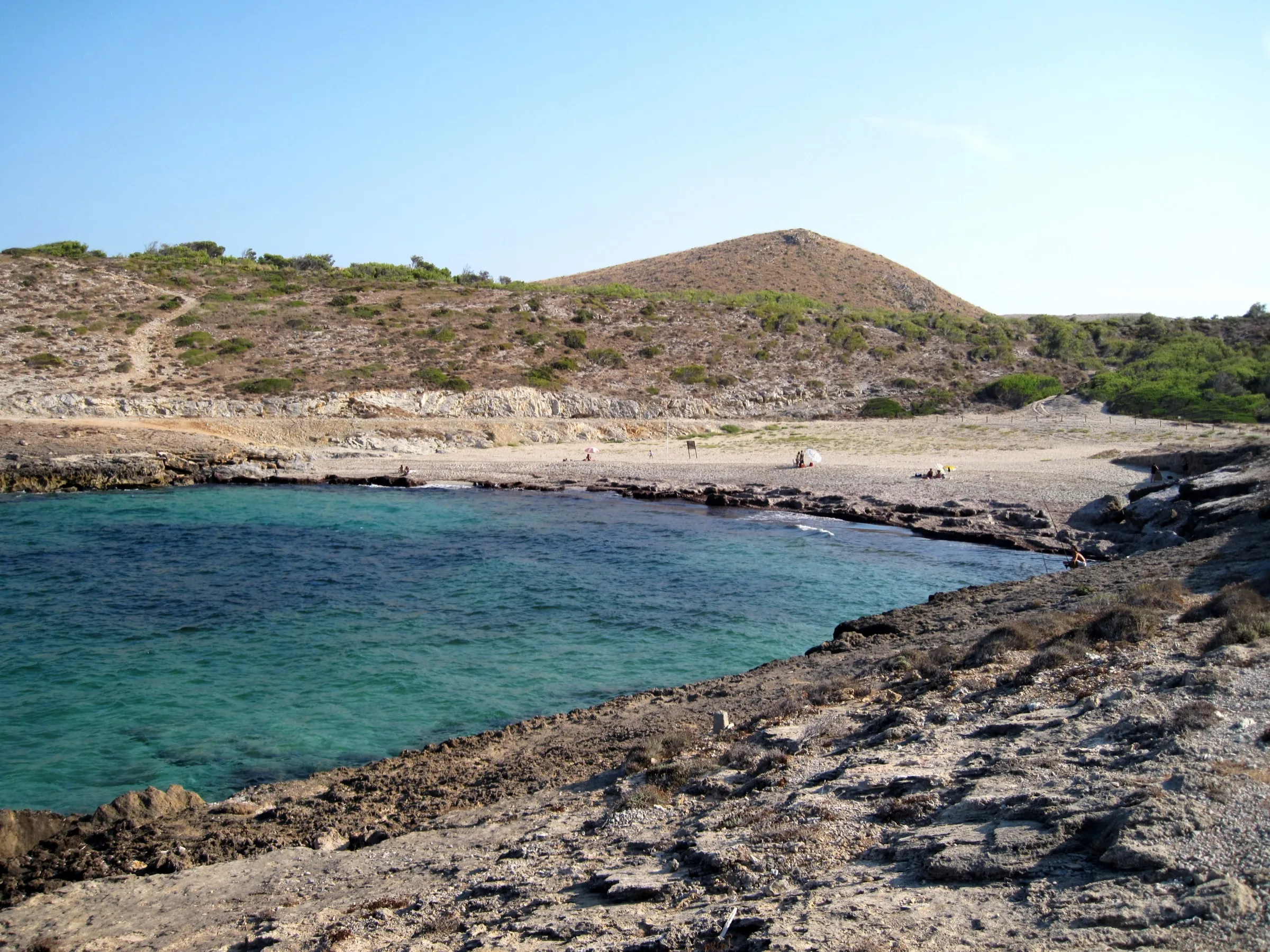 El agua verde y turquesa de Cala Mitjana entre roca plana, con la arena y un monte de matorral detrás
