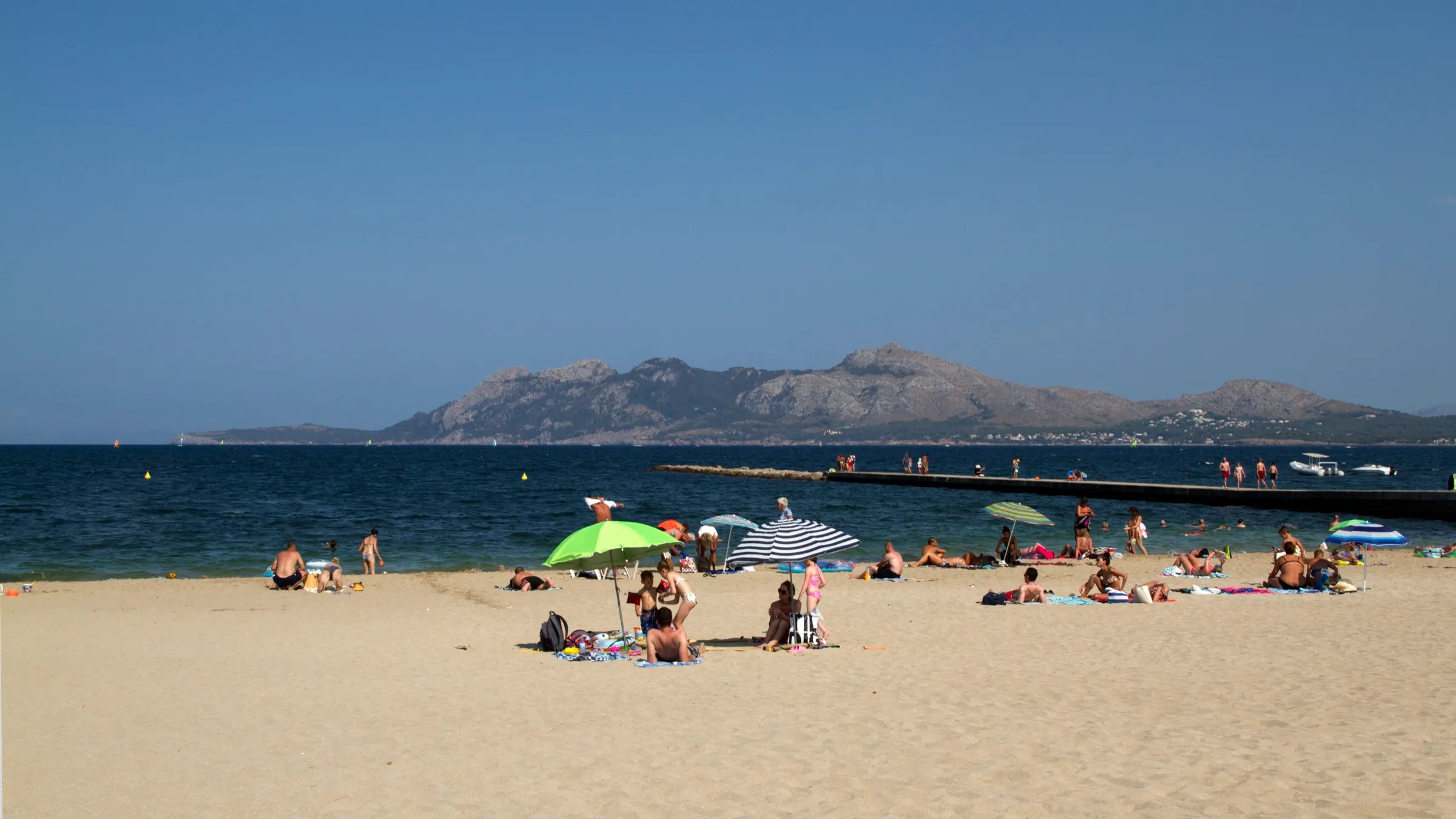 Badegäste mit Sonnenschirmen am Strand von Llenaire, im Hintergrund ein Steg und die Küste der Bucht
