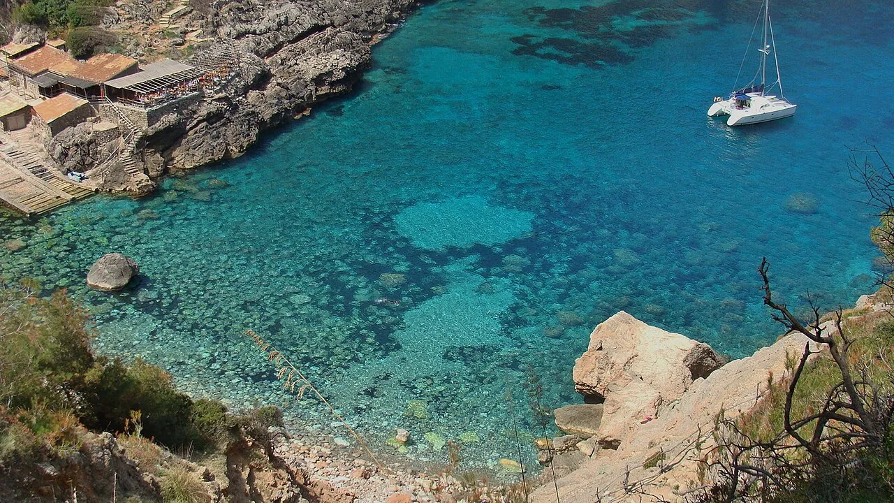 Agua turquesa y transparente de Cala Deià vista desde arriba, con un catamarán fondeado