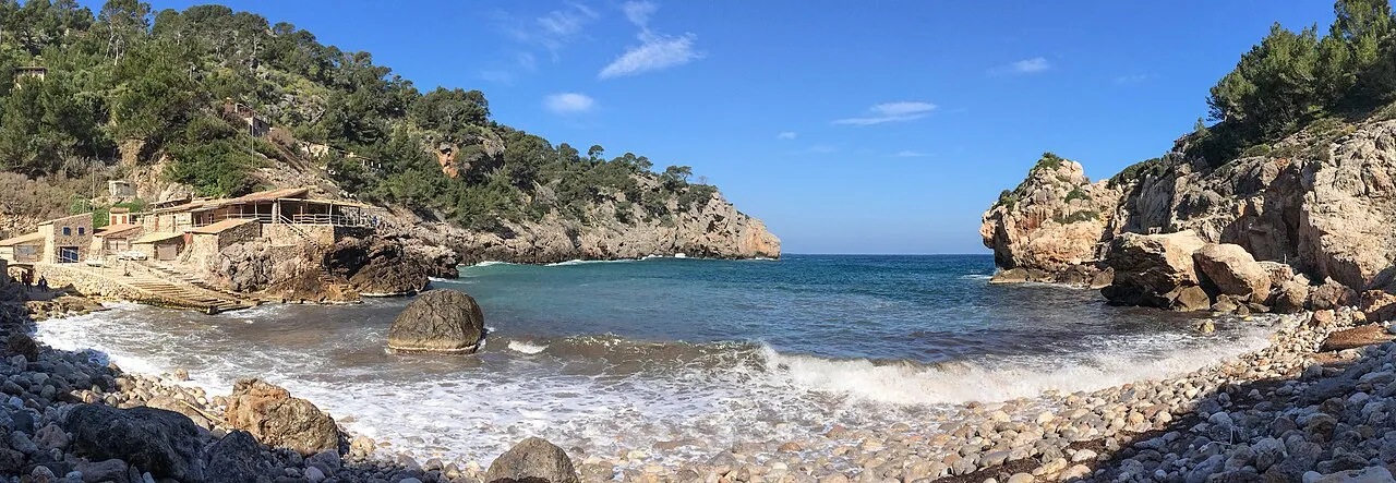 Panorámica de Cala Deià desde la orilla de cantos, con las casetas a la izquierda y la boca de la cala al fondo