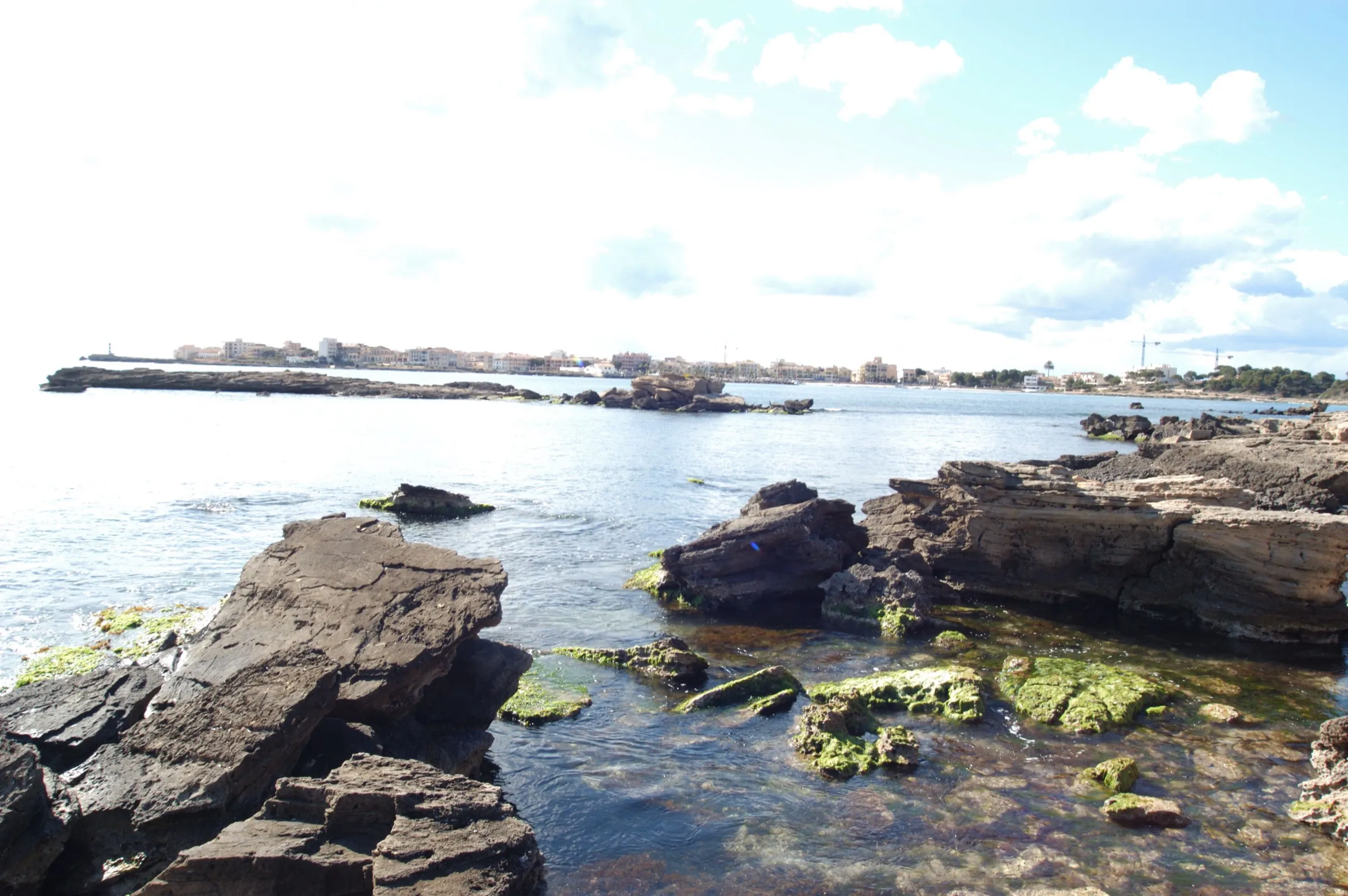 Flache Felsen und klares Wasser bei es Dolç, im Hintergrund der Hafen von Colònia de Sant Jordi
