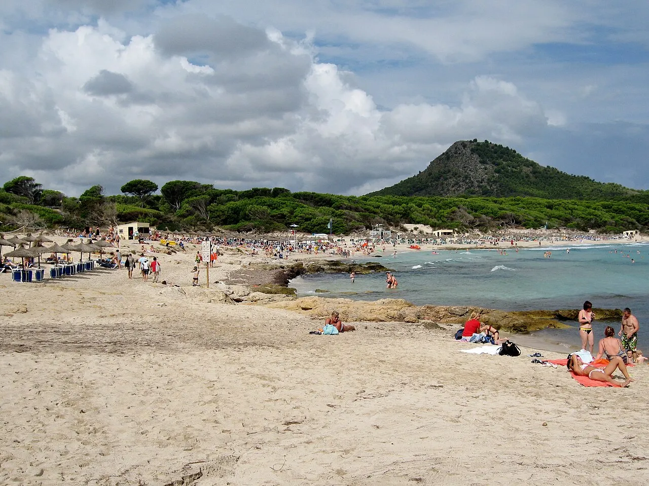 Bañistas en la arena de Cala Agulla, con sombrillas de paja y el cerro de pinar al fondo