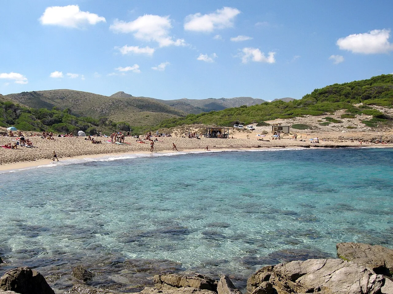 Agua transparente sobre el fondo de arena y roca, con la playa y los montes de Artà al fondo