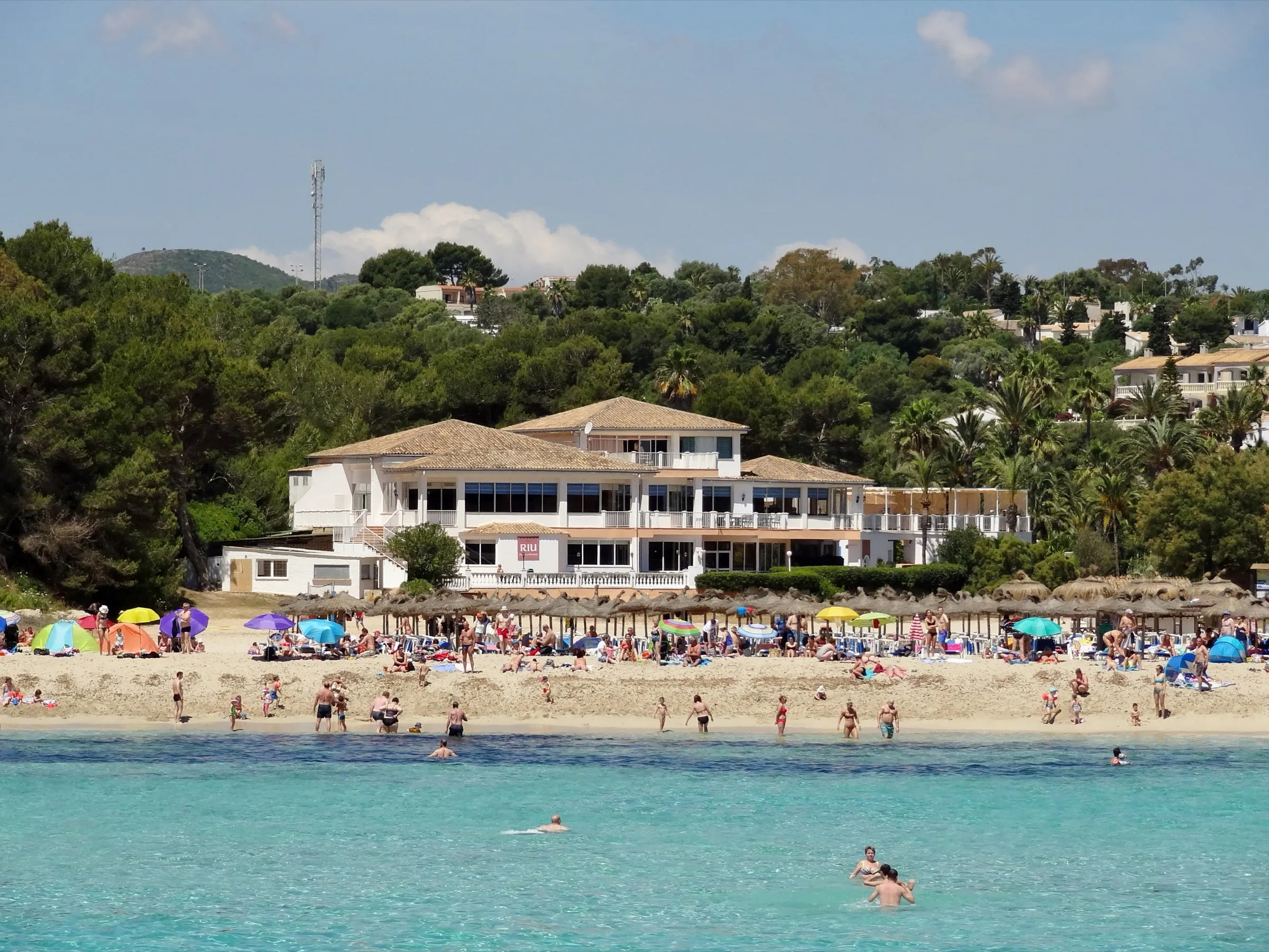 Bañistas y sombrillas en la arena de s'Estany d'en Mas, con un edificio blanco detrás de la playa