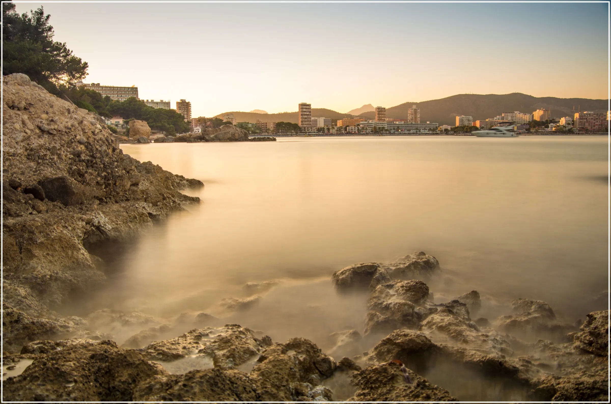 Rocas de la orilla de Cala Vinyes al atardecer, con la costa urbanizada de la bahía al fondo