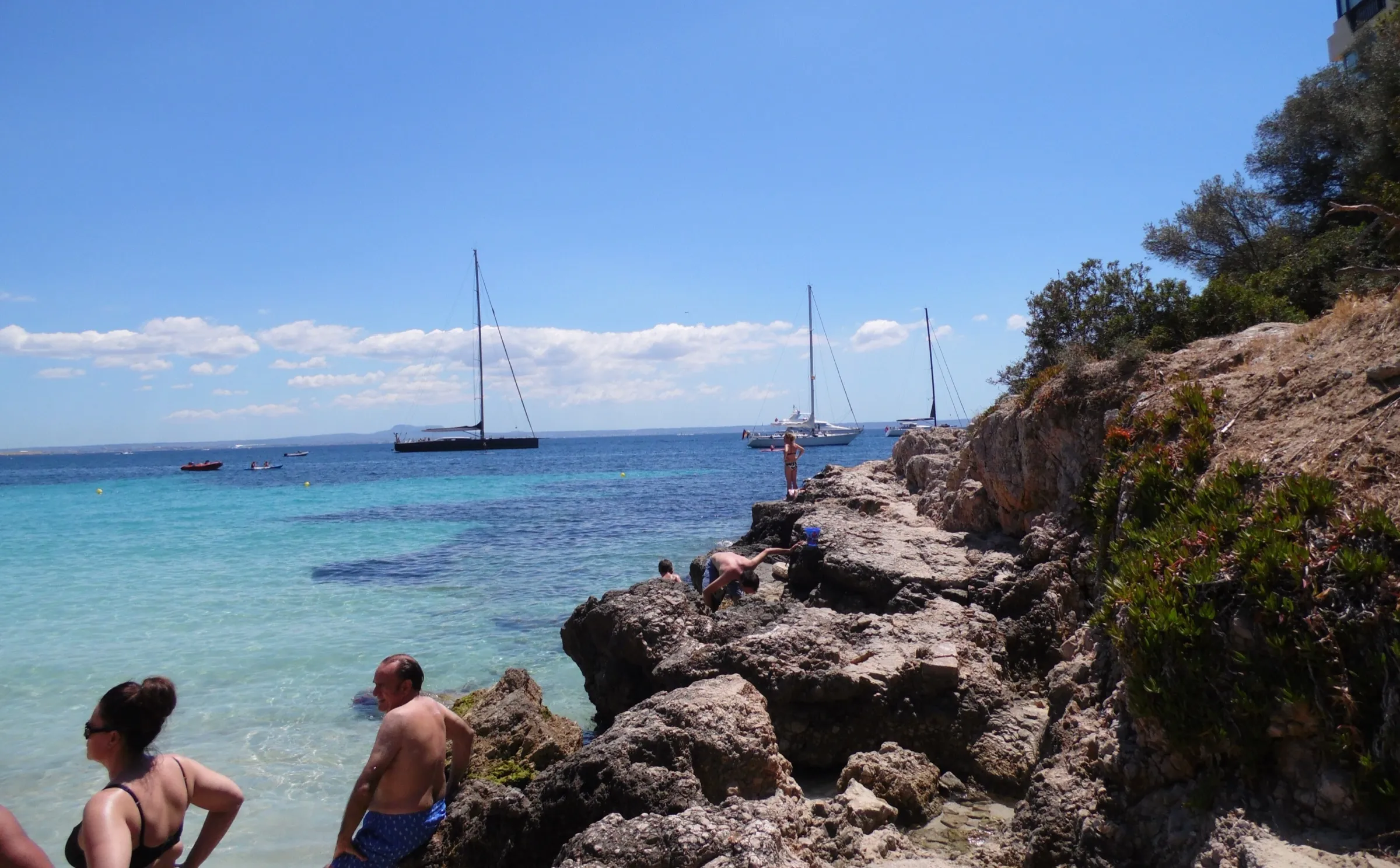 Bañistas junto a las rocas de un lado de la playa, con veleros fondeados en el agua clara