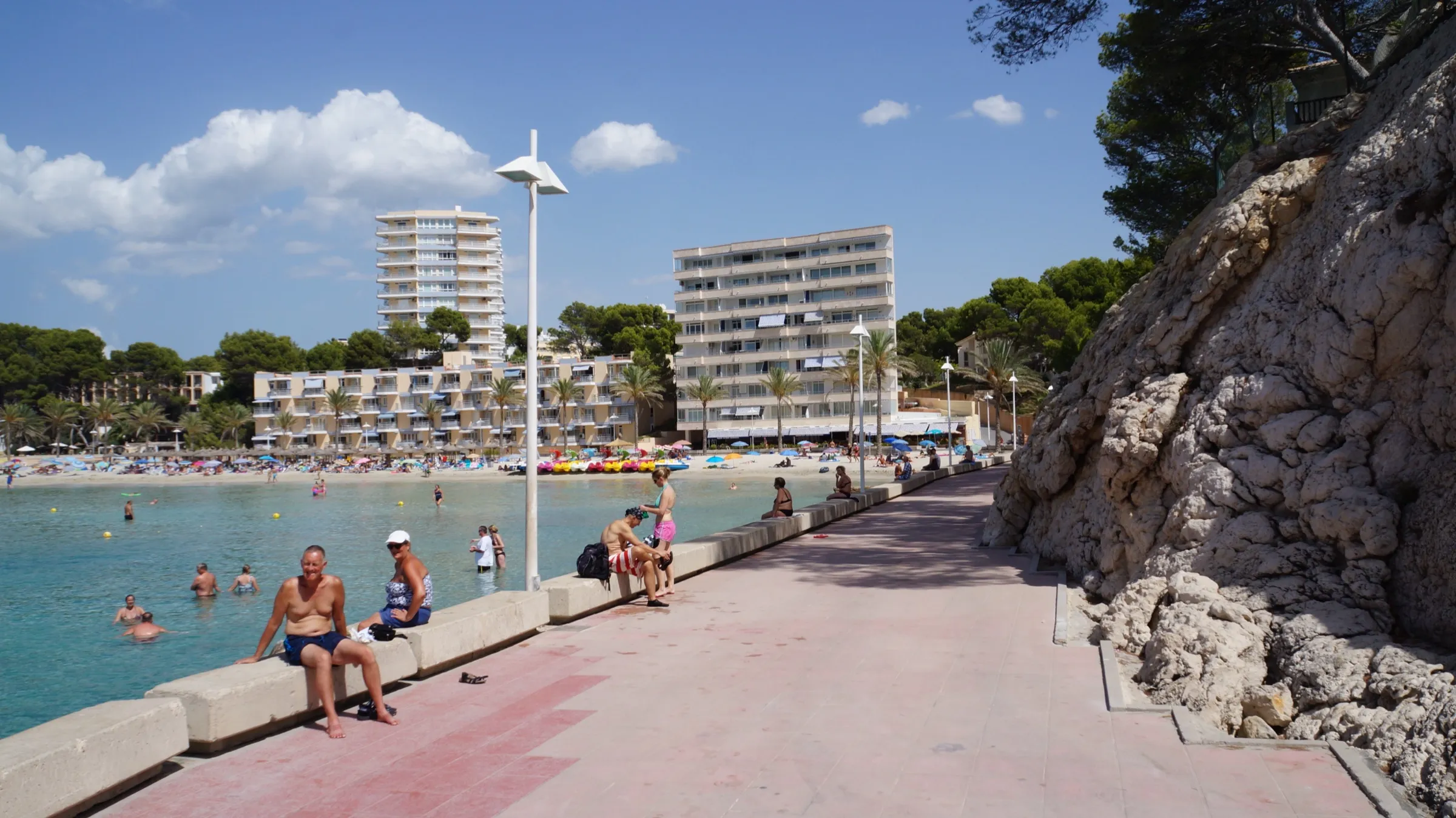 Die Promenade am Fels in Cala Major, mit Badenden auf der Mauer und dem Strand und den Gebäuden im Hintergrund