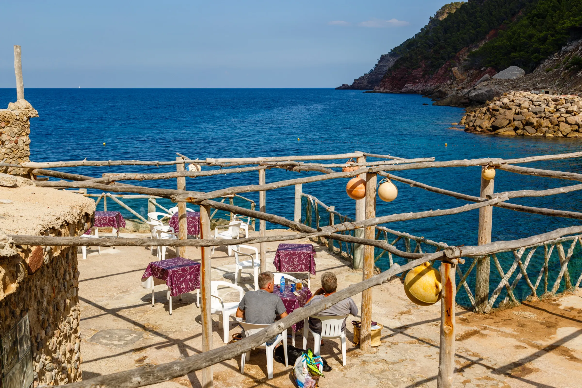 La terraza del bar de Cala d'Estellencs, con barandilla de troncos sobre el agua azul y el espigón de piedras