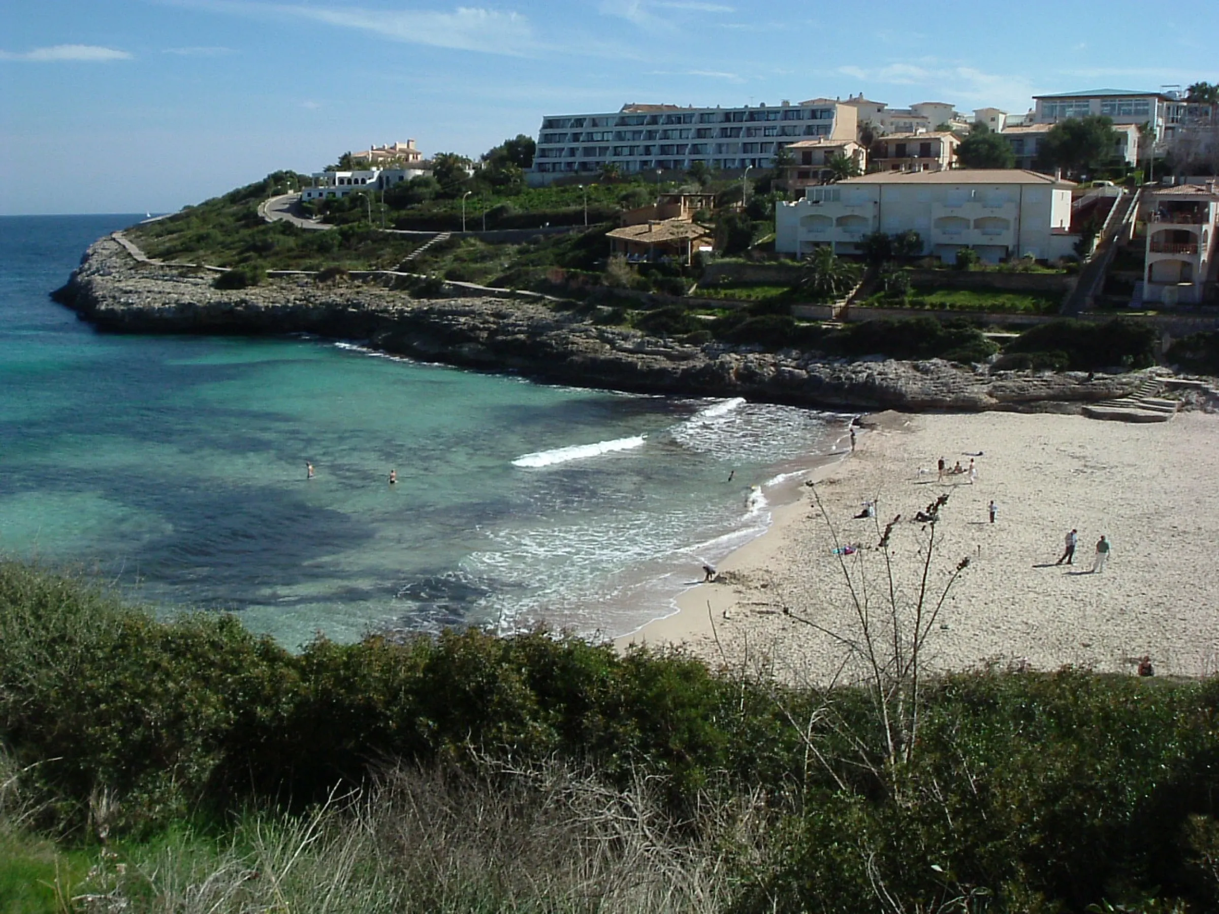 La arena de Cala Mendia fuera de temporada, con la orilla transparente y los edificios de la urbanización sobre la roca