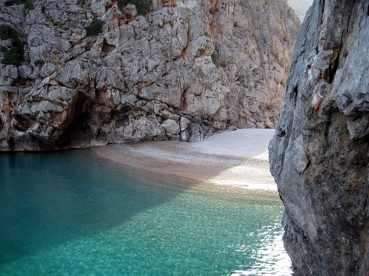 Agua turquesa remansada en la boca del Torrent de Pareis, junto a la orilla de cantos y bajo la pared de roca