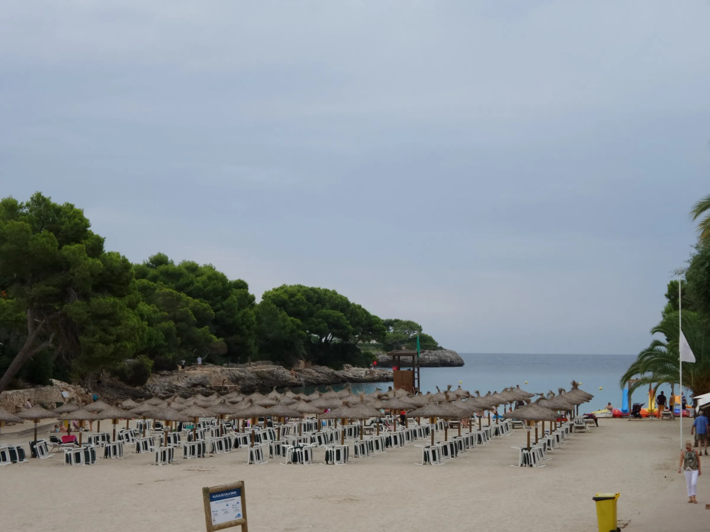 Liegen und Strohschirme auf dem Sand der Cala Gran, dahinter die Buchteinfahrt und das Meer